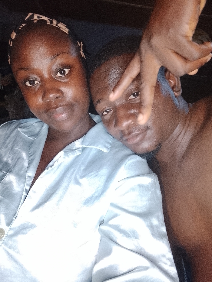 The image captures a close-up selfie of two individuals, a woman and a man, positioned closely together, likely indoors and in a relaxed setting such as a bed or couch. The lighting is dim, suggesting it might be nighttime or a low-light environment.
The woman is on the left side of the frame, facing slightly towards the camera. She has dark skin and is wearing a patterned headband that appears to be brown and light-colored. Her hair is pulled back by the headband. She is wearing a light-colored collared shirt, possibly made of a satin or silk-like material due to its sheen, which appears to be light blue or grey. Her expression is calm, with a slight hint of a smile, and she is looking directly into the camera. Her skin has a natural, slightly dewy appearance.
The man is on the right side of the frame, positioned next to the woman, with his head tilted slightly. He also has dark skin and short, dark hair. He has some facial hair, appearing as stubble or a short beard on his chin and jawline. He is wearing a light-colored collared shirt, similar in style and material to the woman's, suggesting it might be a matching set or a shared garment. His right hand is raised, making a peace sign (V-sign) with his index and middle fingers, partially obscuring the upper part of his face. His expression is also calm, with a subtle smile, and he is looking towards the camera through the gap in his fingers.
The intimate proximity of the two individuals suggests a close relationship. The focus is entirely on their faces and upper torsos. No discernible background elements are visible beyond a dark, out-of-focus area behind them. There is no visible text in the image.