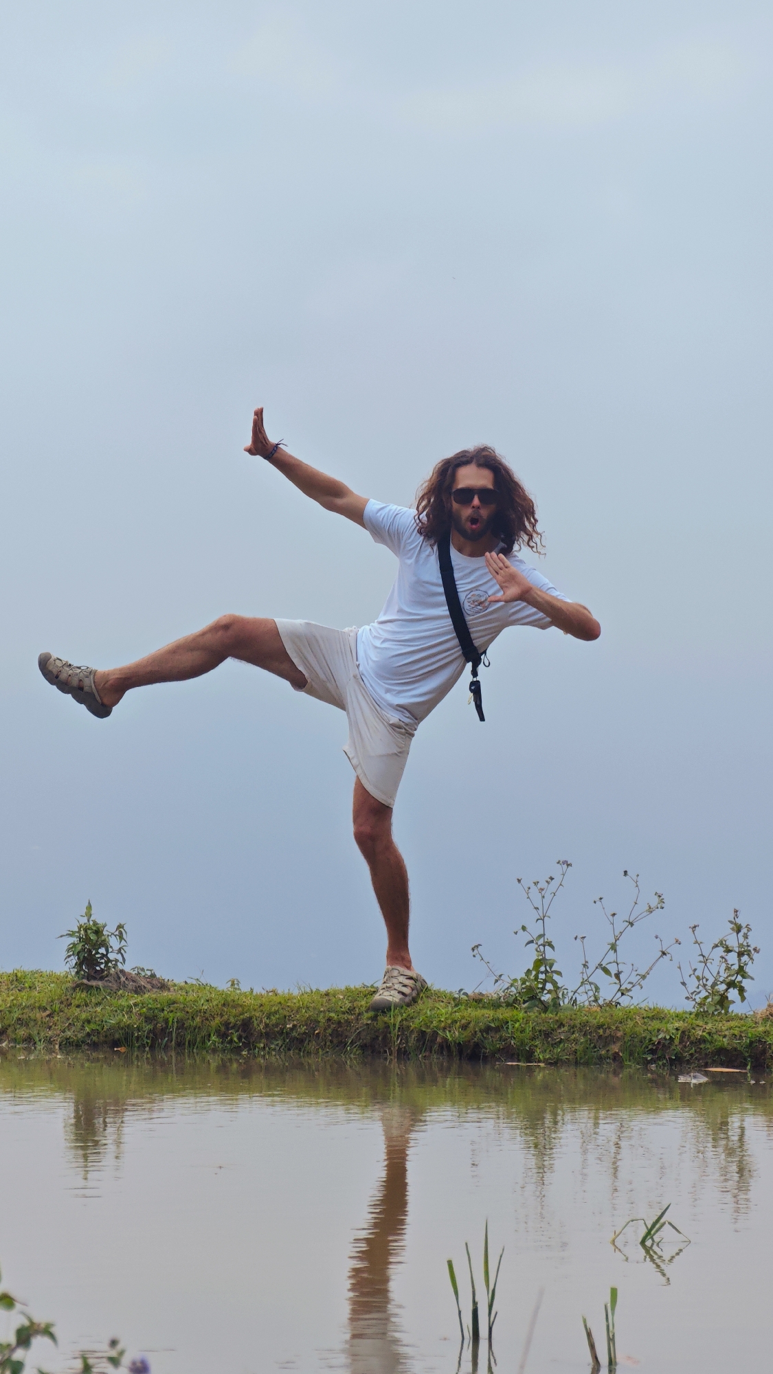 A man with long, wavy brown hair, wearing dark sunglasses, a white t-shirt, and light-colored shorts, strikes a dynamic and playful pose, balancing on one leg. His left leg is extended upwards in a high kick, while his right leg bears his weight. His arms are also dramatically spread, one bent with a flat palm facing forward, and the other extended with fingers splayed, evoking a martial arts or dance move. His mouth is open, suggesting a shout or exclamation. A dark strap, possibly from a bag or camera, crosses his chest.
He stands on a narrow, grassy bank bordering a still body of water, which perfectly reflects his lower body and the surrounding environment. This setting is characteristic of rice paddies in Nậm Dịch Commune, Vietnam. Small green plants are visible along the grassy edge and subtly within the water. The background is a vast, uniformly light grey, overcast sky, providing a clean, ethereal backdrop that isolates the subject. The diffused light indicates daytime. The image captures a moment of energetic expression and balance within a tranquil, agricultural landscape. No text is visible.