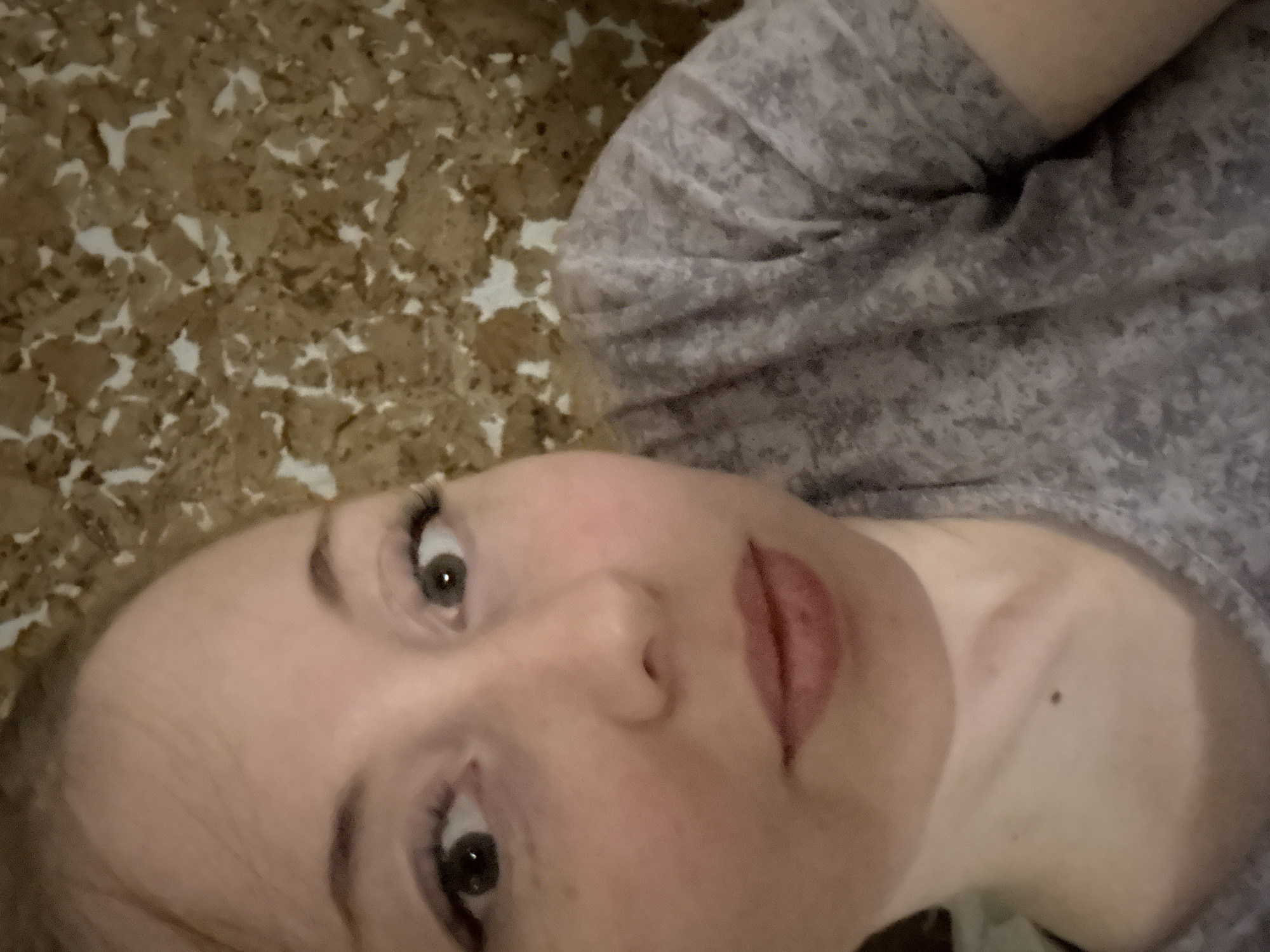 A close-up, angled selfie shows a person with fair skin and light hair. The person is wearing a grey, patterned top. Their face is the primary focus, with prominent eyes, nose, and lips. The background features a brown and white speckled surface. The location and city are unknown.