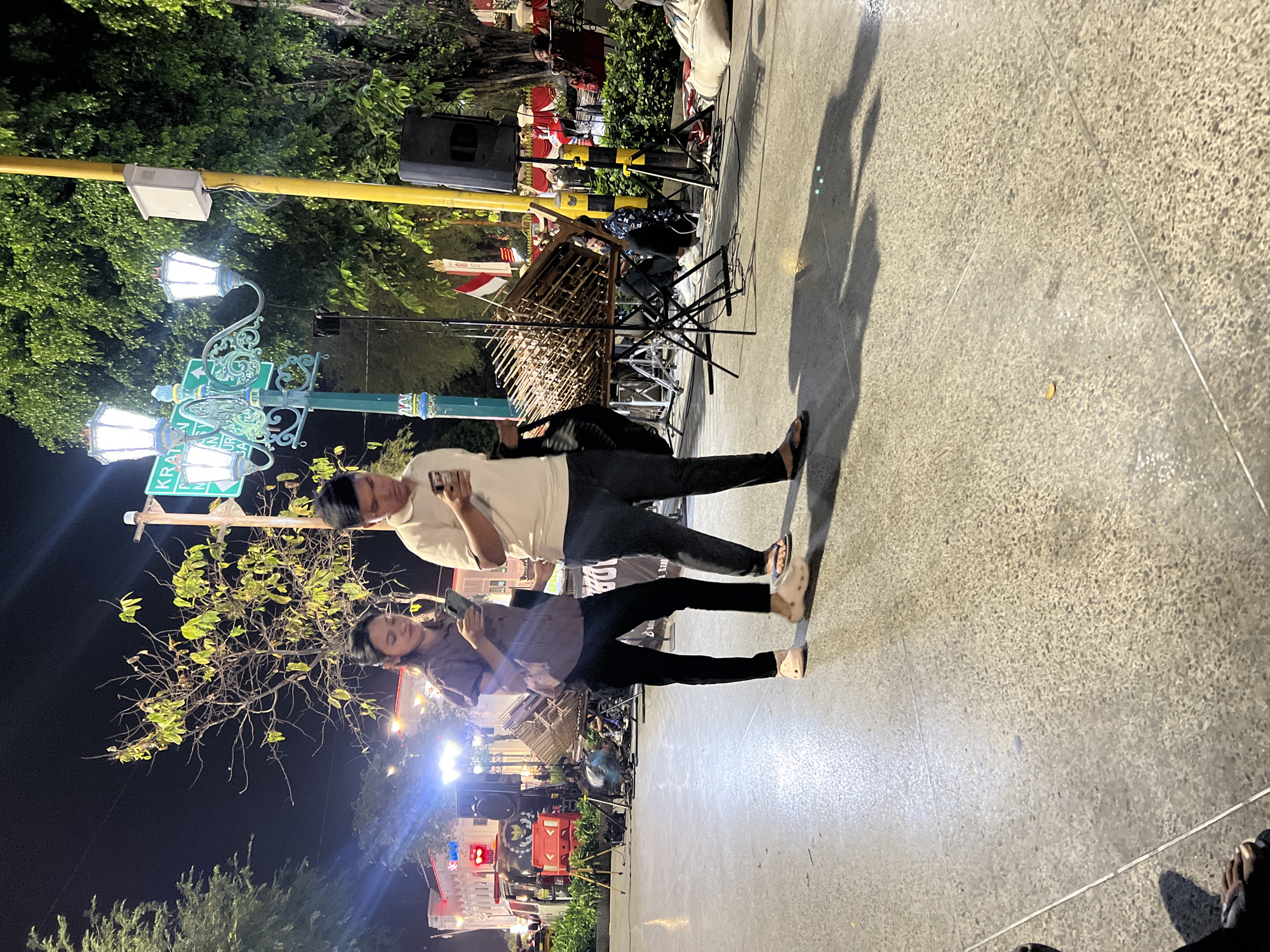 An evening scene shows a young man and woman walking on a paved outdoor area, both looking at their phones. The man is wearing a light-colored polo shirt and dark pants, while the woman is in a striped purple shirt and black pants. They are walking on a textured, gray pavement that reflects some light. Behind them, a traditional Indonesian musical instrument, possibly angklung, is set up on stands with microphones. A street lamp with ornate detailing and multiple lights is visible, along with a street sign reading "KRATON" and directional arrows. Trees with lush green foliage frame the background. The area appears to be a public space in the city of Yogyakarta, Indonesia, possibly near the Kraton (Sultan's Palace) area, which is known for its cultural performances and evening activities. There are also stage lights, speakers, and some people seated in the background, suggesting an ongoing event or performance.