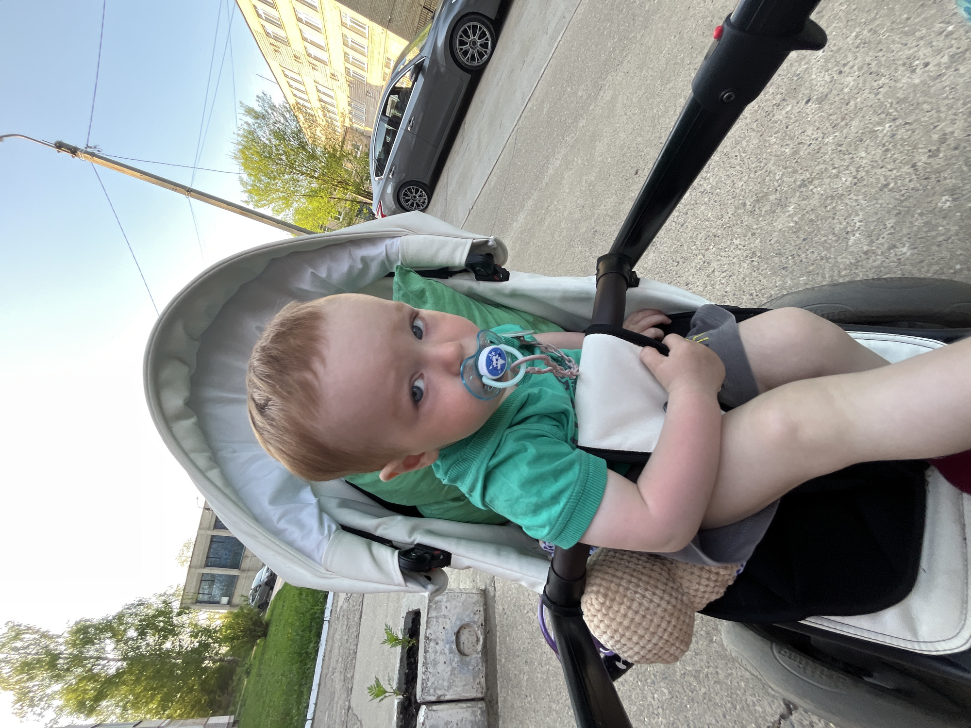 A baby with fair skin and light reddish-blonde hair is sitting in a stroller, facing the camera with a pacifier in their mouth. The baby is wearing a green shirt and is holding a white and grey blanket. The stroller is white with black frame elements and wheels, and it appears to be on a sidewalk.
The surrounding environment includes a paved road with a parked grey car, a utility pole, and buildings. Lush green trees and lawn are visible in the background, along with a concrete block with small plants growing out of a cutout.
Given the clues, the location is likely in a residential or urban area in Chaikovsky, Russia, with some greenery near the pavement.