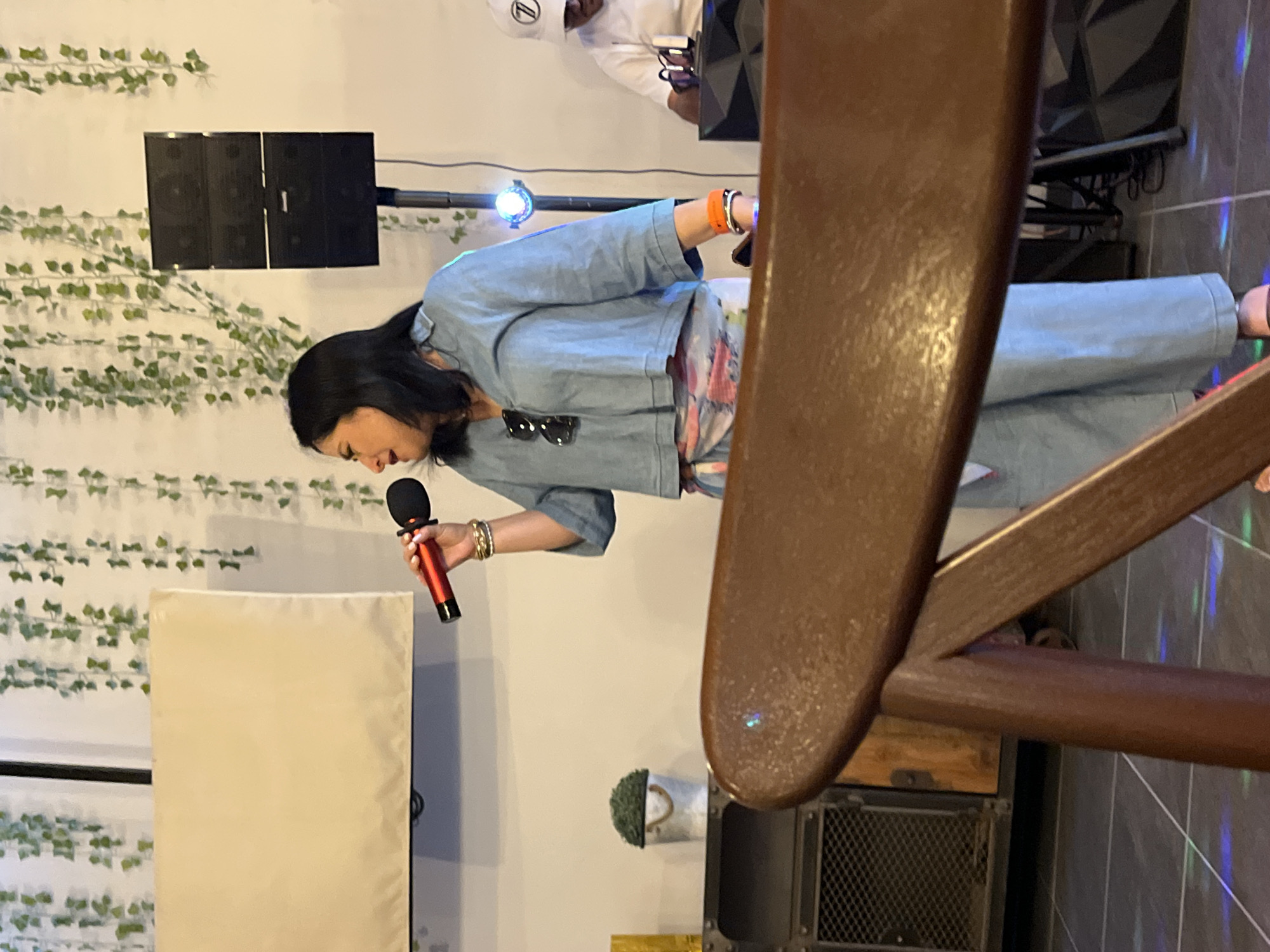A woman with short black hair is holding a microphone and singing. She is wearing a denim jacket, a colorful top, and denim pants. Behind her is a wall with green vines and a speaker system. There is a white object to her left. In the foreground, there is a brown wooden chair. The scene appears to be indoors, potentially at a karaoke bar or a private event. The location is San Bernardino Tlaxcalancingo, Mexico.