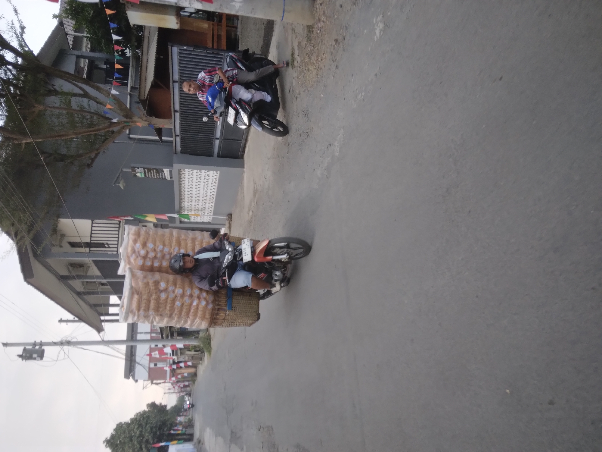 In the city of Desa Pamijen, Kecamatan Sokaraja, Indonesia, two men are riding motorcycles on a paved road. The man in the foreground is on an orange and black motorcycle, carrying a large, stacked basket of snacks behind him, secured to the back of his vehicle. He is wearing a helmet, a dark jacket, and shorts. The motorcycle has a license plate that reads "B 4410 NP". To his right, another man on a black motorcycle is wearing a red and white checkered shirt and gray pants, also riding on the road. The environment includes residential buildings, trees, and utility poles. The sky is overcast.