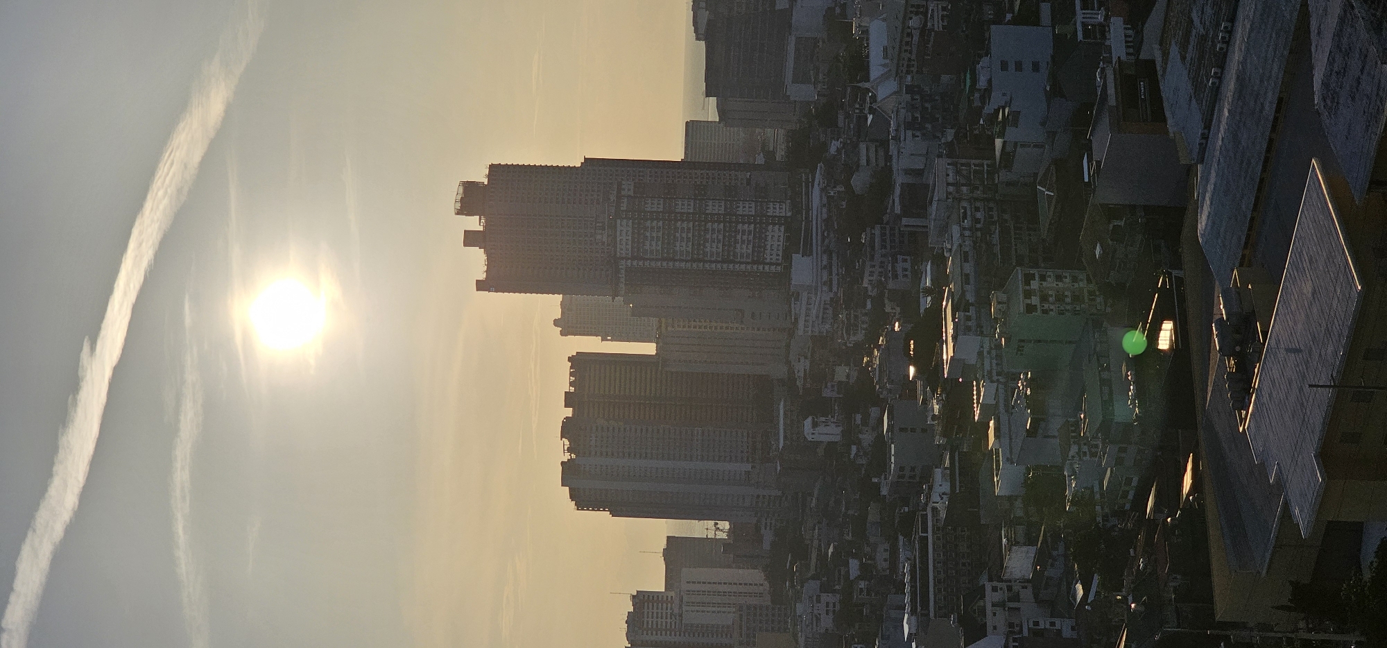 This is a vertical, high-angle shot of the cityscape of Makati, Philippines, during what appears to be late afternoon or early morning, given the low angle of the sun and the warm, hazy light. The sun, a bright white orb, is positioned high in the sky, partially obscured by a thin layer of clouds. A prominent contrail, a long, white streak left by an aircraft, cuts diagonally across the sky from the upper left to the middle right.
Below the sky, a dense urban landscape unfolds. Numerous buildings of varying heights dominate the scene. Tall, modern skyscrapers, some appearing to be under construction with visible scaffolding, stand clustered in the middle ground. These structures are characterized by their repetitive window patterns and flat roofs. In the foreground and middle distance, a more varied collection of lower-rise buildings, including apartment complexes and residential structures, create a textured urban fabric. Many of these buildings have visible rooftops with air conditioning units, water tanks, and other utilitarian features. The overall color palette is muted, with shades of gray, white, and beige prevailing, accented by the warm, golden light of the sun. Shadows are cast by the buildings, highlighting the depth and dimensionality of the cityscape. No people or vehicles are clearly discernible in the frame. The environment is clearly urban, characterized by dense construction and infrastructure.