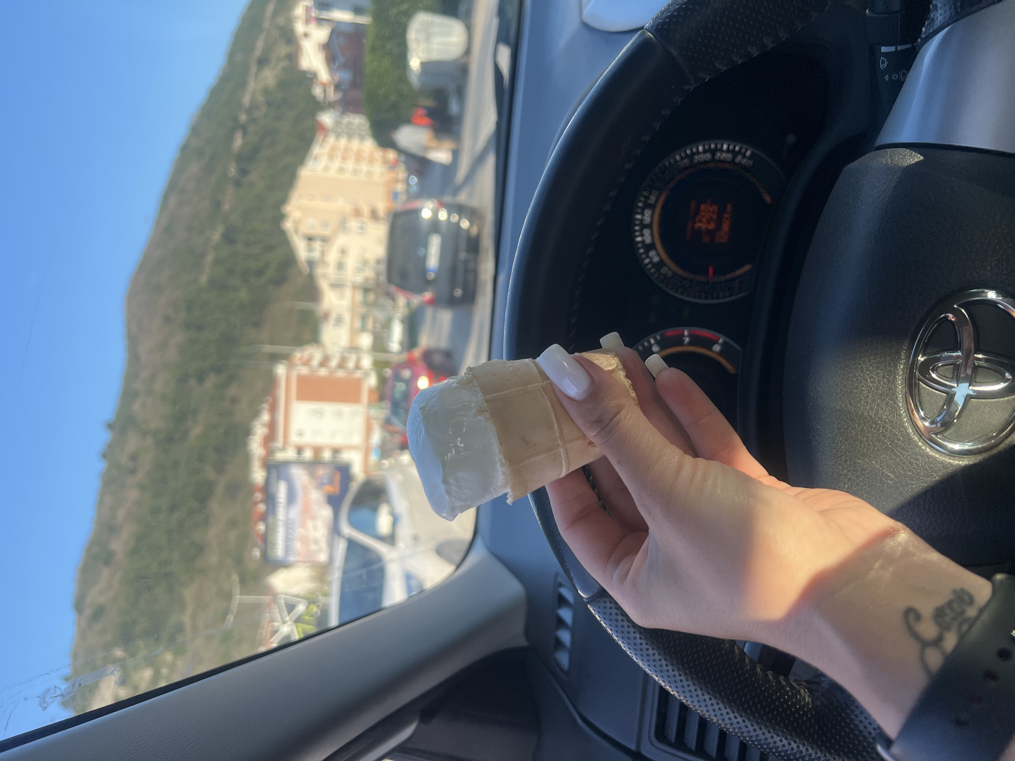A person is holding a single scoop of white ice cream in a waffle cone while seated in a car. The driver’s side dashboard and steering wheel are visible. Outside the car window, there is a street lined with buildings. A large, green, rocky hill occupies the background. The person has a tattoo on their wrist and is wearing a black watch band. The car is on the road near Jadranski put in Budva, Montenegro.