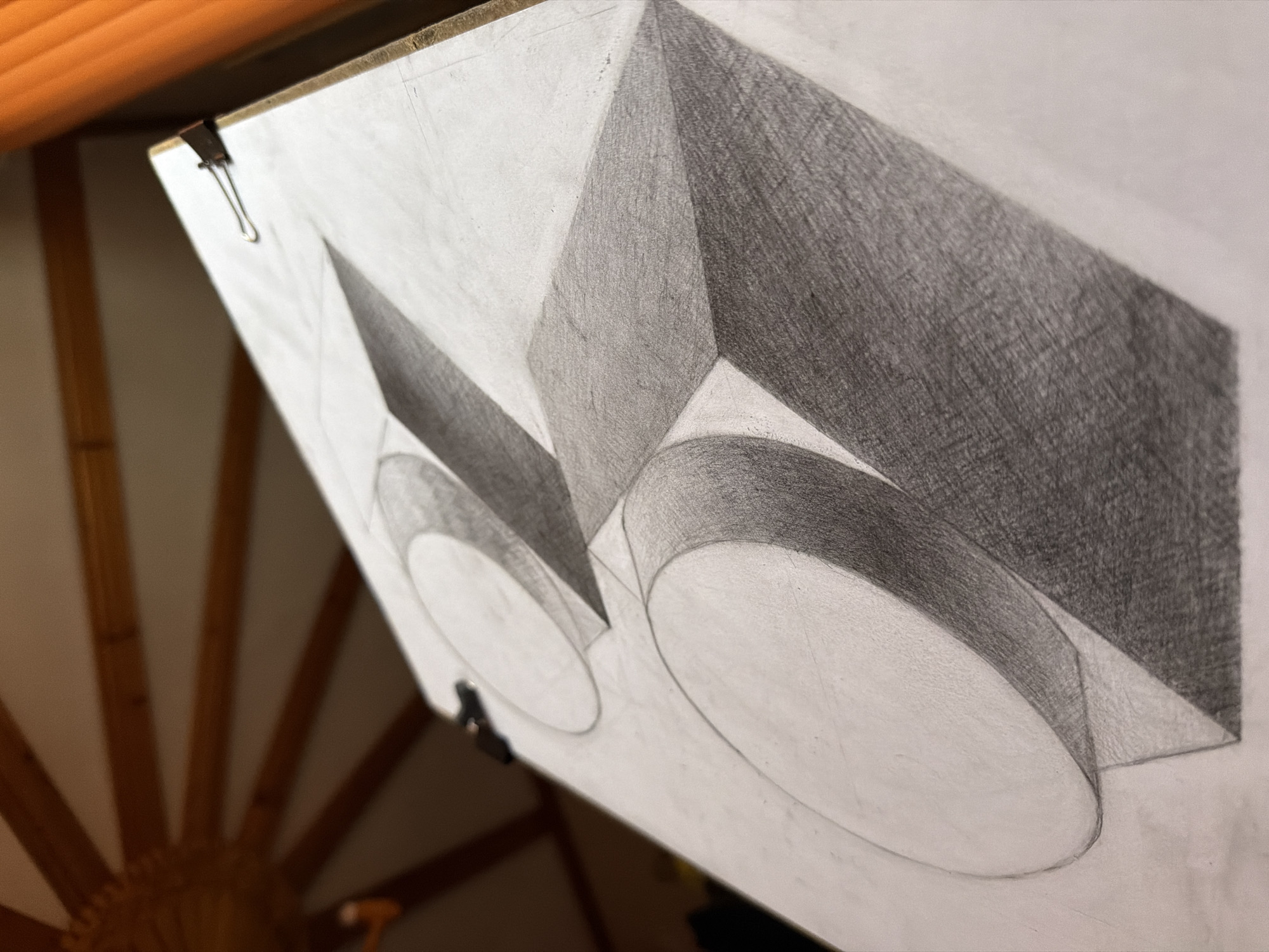 The image depicts a drawing of geometric shapes on a sheet of paper held by clips, tilted at an angle. The shapes consist of two overlapping circles and rectangular prisms. One prism is lightly shaded, while the other is heavily shaded with graphite. The setting appears to be indoors, with a wooden structure with radiating beams visible in the background. There is no information to suggest that the location is in the city of Velingrad, Bulgaria.