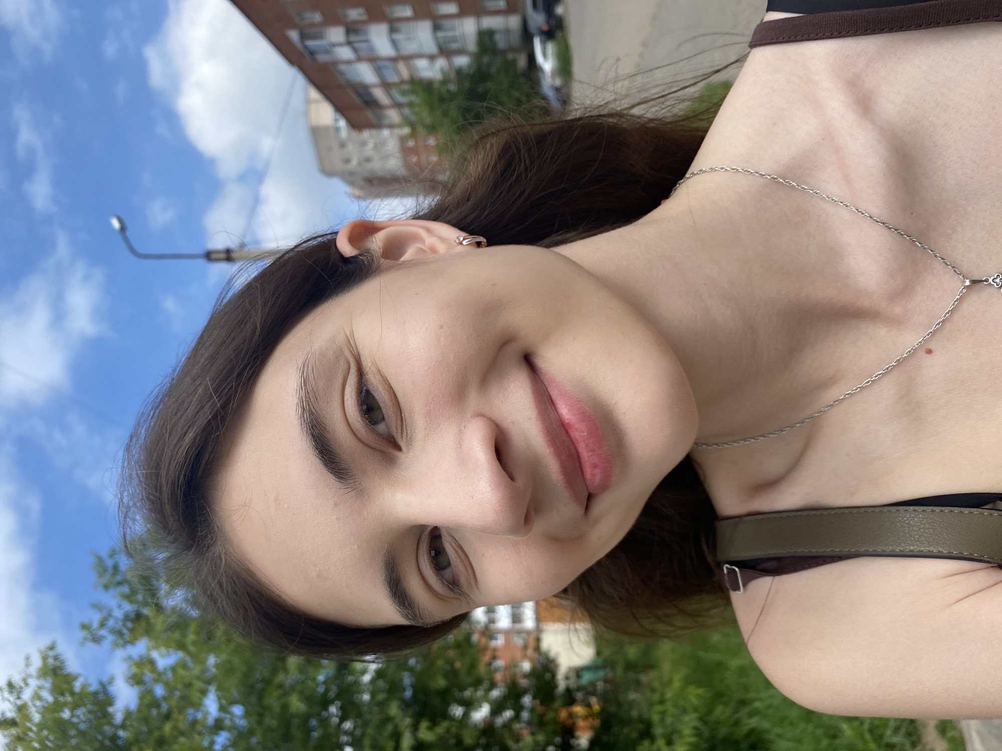 A young woman with long, dark hair and fair skin smiles at the camera. She is wearing a silver necklace and a small hoop earring in her left ear. She is outdoors in a city environment. Behind her, trees and apartment buildings are visible. The sky is blue with white clouds. The image appears to have been taken on a sunny day. The location is Votkinsk, Russia.