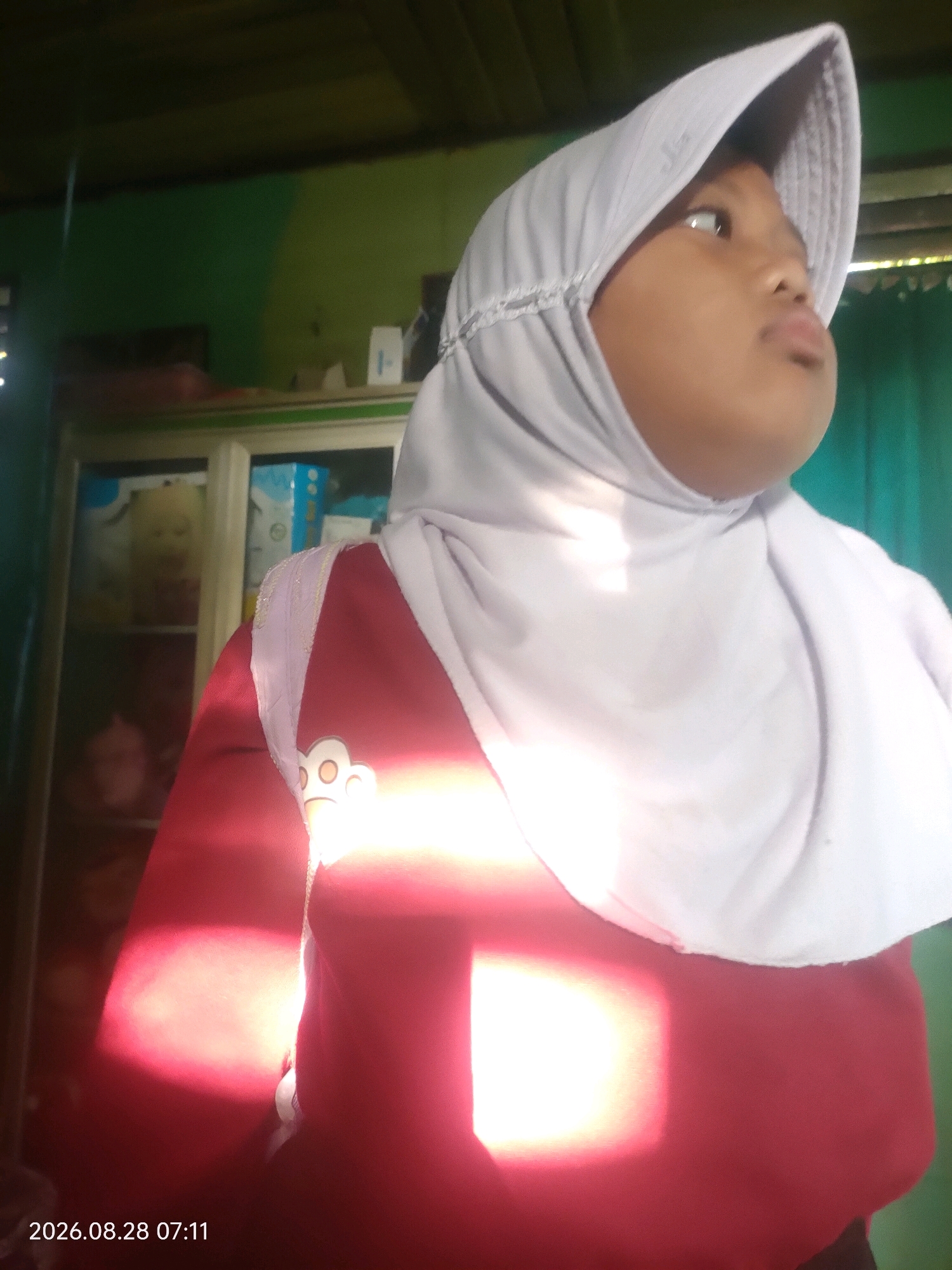 A young girl, wearing a light gray hijab and a dark red shirt with a cartoon paw print emblem on the chest, is shown in a medium-close-up shot. She is looking upwards and to the right, her expression neutral. The background consists of a green wall and a cabinet with several shelves containing various items, including what appears to be stuffed animals or dolls. A timestamp "2026.08.28 07:11" is visible in the bottom left corner of the image. The scene suggests an indoor setting, potentially a home or a room within a building. No specific details about the city of Kuaro, Indonesia are discernible from the image itself, as the location information is provided as context for the description and not visually present within the frame.