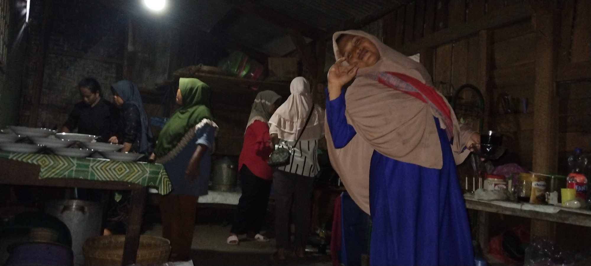 The image captures a dimly lit interior, likely a home or community gathering space, in Tanjung Ratu Ilir, Indonesia. In the foreground, a woman wearing a light brown hijab and a blue dress poses for the camera, holding up a hand with an extended index finger and thumb. To her left, a group of people are gathered around a table laden with white plates. One woman, wearing a green hijab and a blue and brown outfit, stands facing left, appearing to be involved in food preparation or serving. Another woman in a dark blue headscarf and black top is also at the table. In the background, a woman in a red top and black pants, and another woman in a light pink hijab and a striped top are visible. The environment appears rustic, with wooden walls and a corrugated metal roof visible. Shelves in the background hold various items, including containers, bottles, and what appears to be a bicycle wheel. The overall scene suggests a communal event or meal preparation taking place in a modest setting.