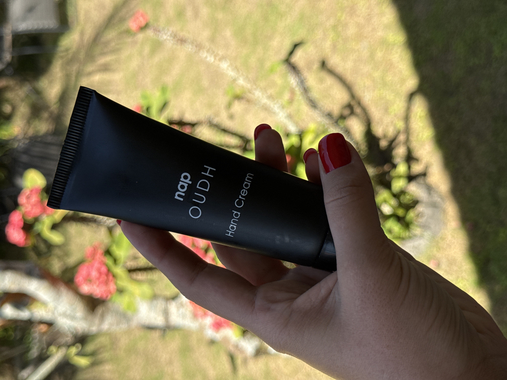 A woman's hand holds a tube of "nap OUDH Hand Cream" against an outdoor background. The tube is matte black with white lettering. The hand has red nail polish. The background appears to be a yard or garden in daylight. The location is Pejeng, Indonesia.
