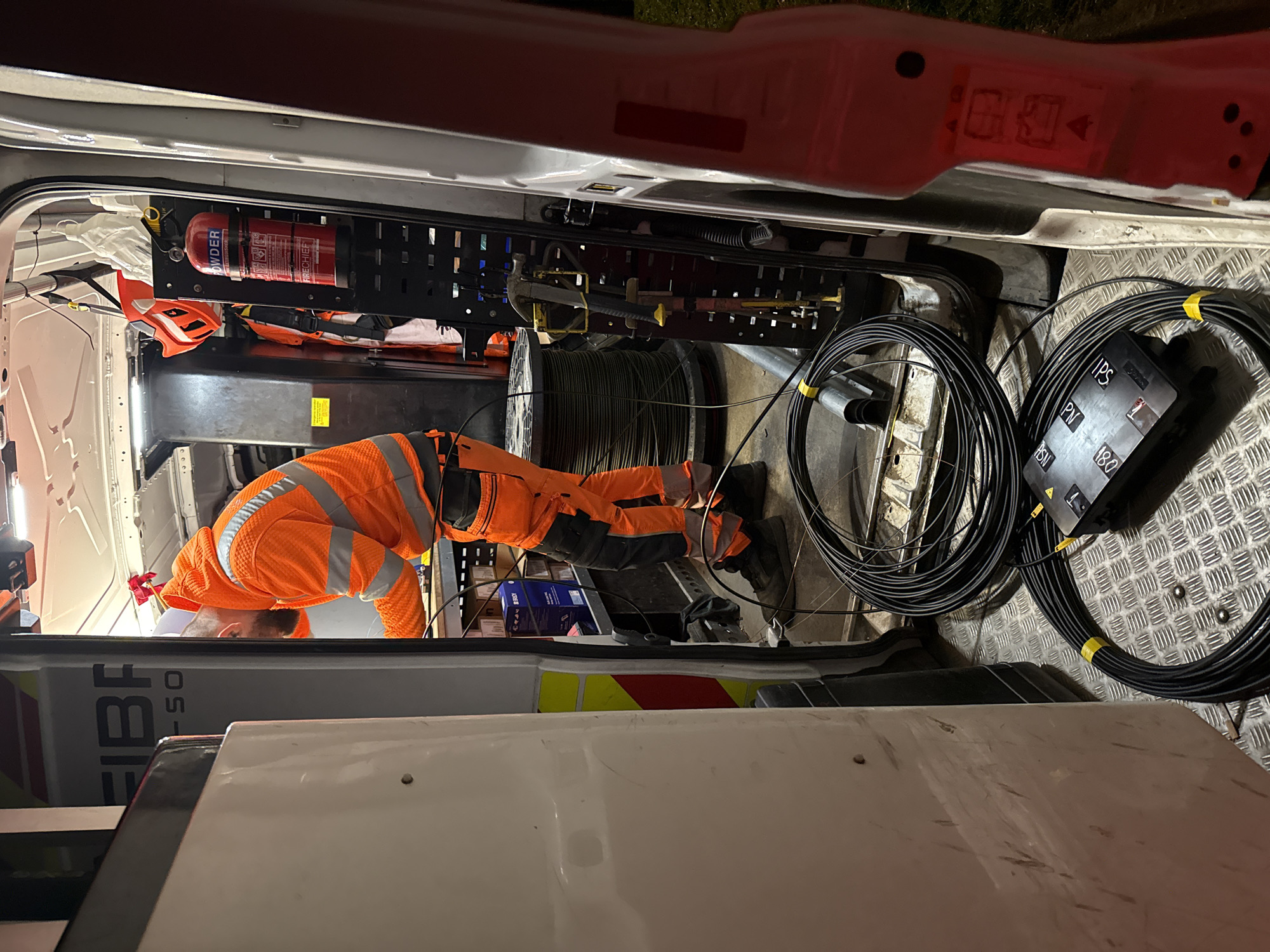 A man wearing an orange high-visibility jacket is bent over, working inside the rear of a van. The interior of the van is equipped with storage racks holding equipment, including a fire extinguisher. A large spool of cable and coils of black fiber optic cable are on the floor of the van. The van appears to be a utility vehicle associated with fiber optic installation, based on the "FIBRE" logo on the side. The location is potentially within Mid Suffolk, United Kingdom.