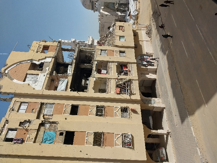 An urban scene in Khan Yunis, Palestinian Territory, depicts a severely damaged multi-story building under a clear blue sky. The beige-colored structure, likely residential, exhibits extensive destruction across multiple floors, with large sections of walls missing, exposing interiors, shattered windows, and displaced debris. Pockmarks, consistent with bullet impacts or shrapnel, are visible across the facade. Laundry hangs from several balconies and windows. In the upper right background, a white dome structure, part of a further extensively damaged building, is visible.
On the paved street in the foreground, at least ten individuals are present. Approximately five to six individuals, dressed in dark uniforms, are scattered across the street and sidewalk; one is walking from left to right on the road, while others are standing or seated on the ground. A group of individuals is gathered on the sidewalk near the entrance of the primary damaged building, where a silver vehicle is parked in a covered ground-level area beneath the building's arches. The overall environment indicates widespread architectural destruction and human presence amidst the rubble.