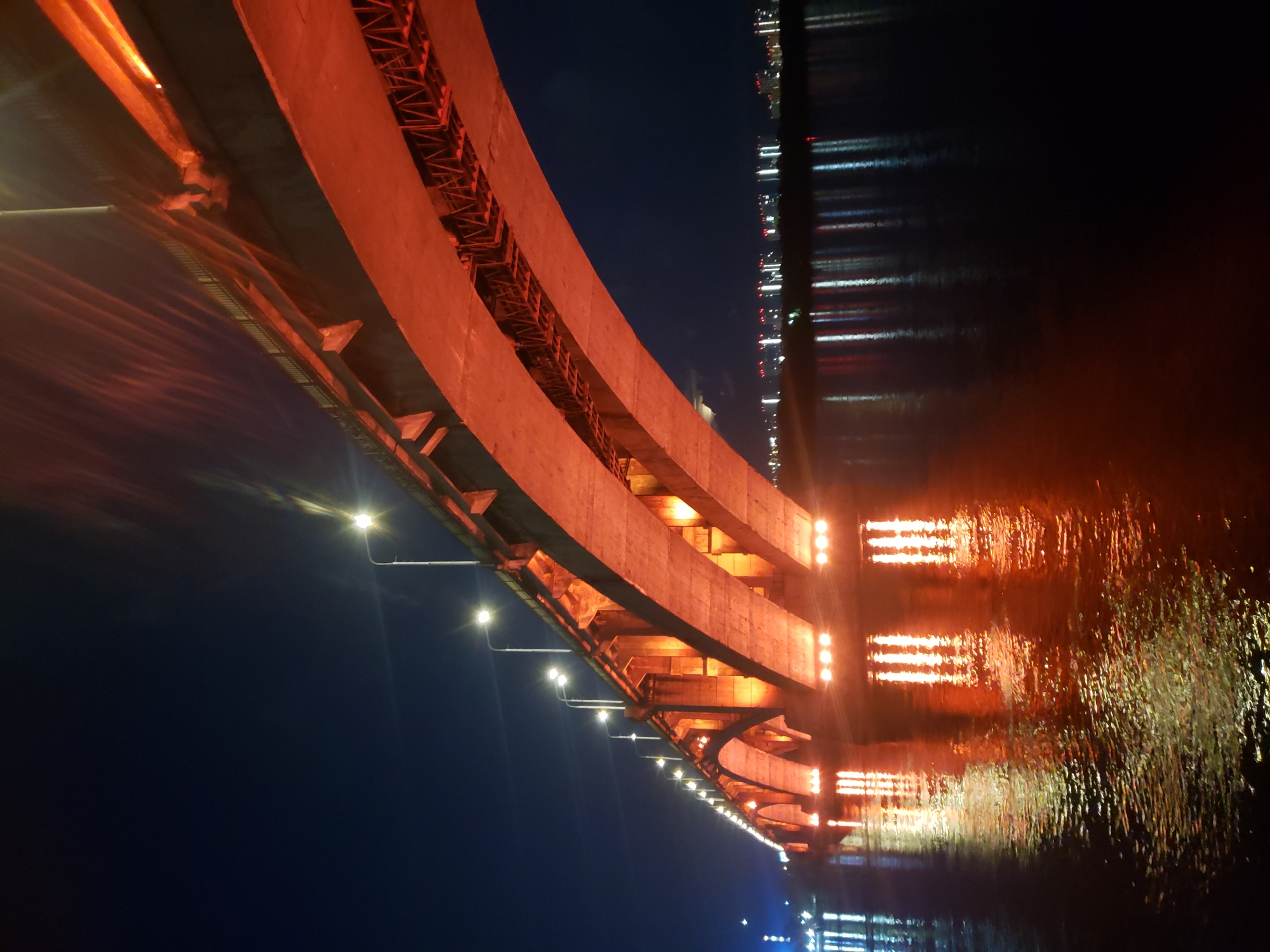 The image is an upward, angled view of a bridge at night. The bridge is lit with warm, orange-toned lights that cast a glow on the underside of its structure. The bridge appears to be made of concrete and metal, with visible beams and supports. Along the edge of the bridge, streetlights are visible, emitting bright white light. Below the bridge, a body of water reflects the lights from the bridge and distant city lights, creating a shimmering, abstract pattern. The water is dark, suggesting a clear night sky. The overall mood is serene and somewhat dramatic, due to the interplay of light and shadow. There is no visible text in the image.