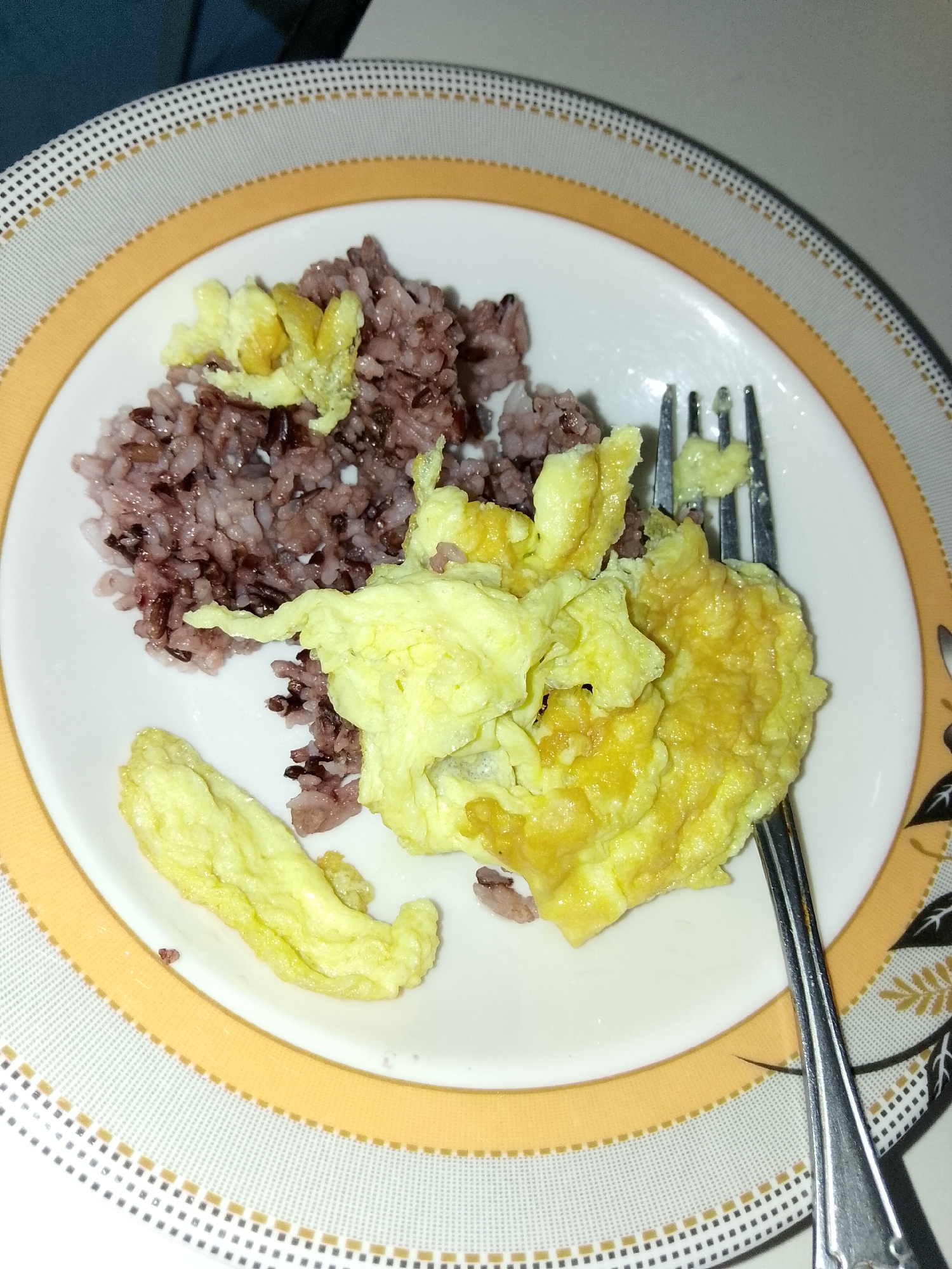 A plate of scrambled eggs with purple rice and a fork is shown on a placemat. The meal is on a white plate with a yellow and black patterned rim, and it sits on a round placemat with a similar design. The lighting suggests an indoor setting, likely a dining area. There are no visible people or specific landmarks that would indicate a location in Davao City. The focus is on the food and the tableware.