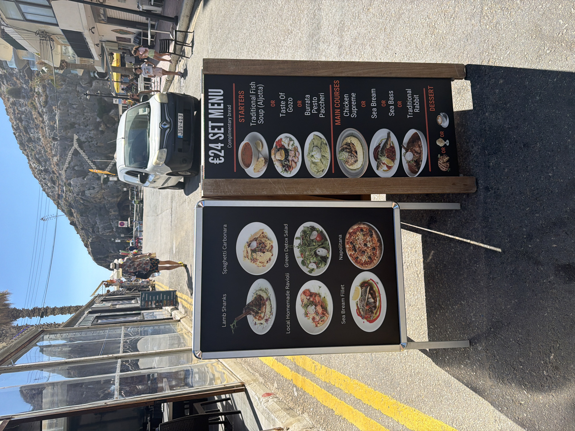 A double-sided A-frame menu board stands on a paved street in Xlendi, Malta. The left side displays images and names of dishes, including "Lamb Shanks," "Spaghetti Carbonara," "Local Homemade Ravioli," "Green Detox Salad," "Sea Bream Fillet," and "Napoletana." The right side advertises a "€24 Set Menu" with options for starters like "Traditional Fish Soup (Aljotta)" and "Taste Of Gozo," main courses such as "Chicken Supreme" and "Sea Bream," and dessert. The street is lined with buildings and a palm tree. In the background, a white van is parked, and several people are visible walking and standing. The environment appears to be a sunny day, with shadows cast on the ground.
