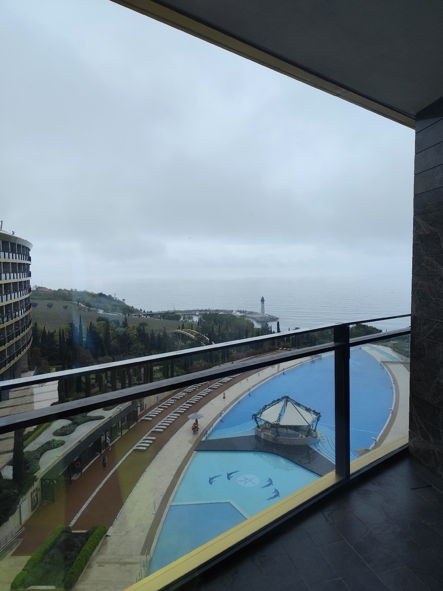View from a balcony overlooking a resort area. The scene includes a large swimming pool with a central island feature, surrounded by lounge chairs and pathways. In the background, there is a coastline with a lighthouse visible on the horizon. The sky is overcast. To the left is a multi-story building. The location appears to be a coastal resort, possibly near a city. The city and country are unknown.