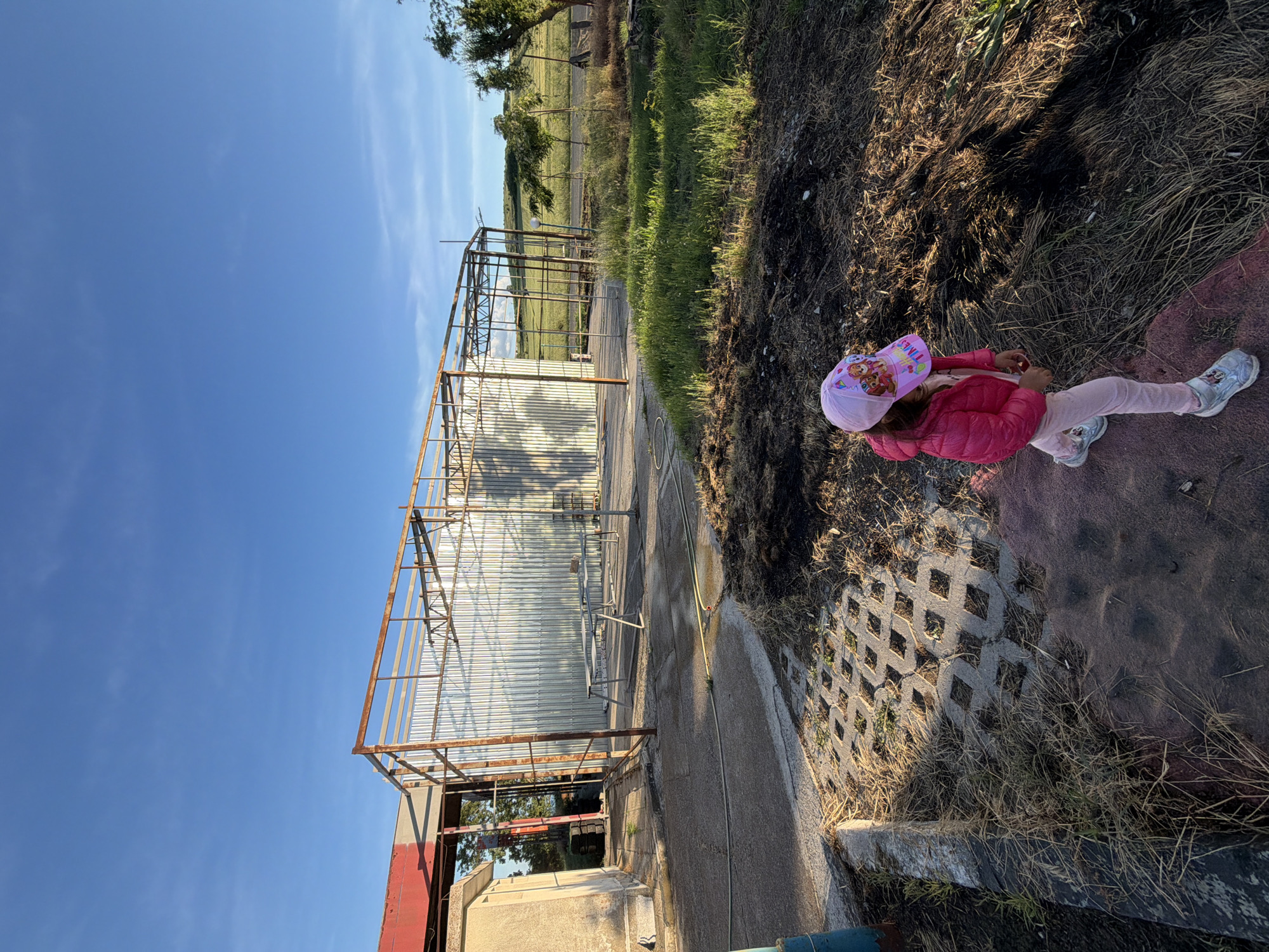 A young girl in a pink jacket, pink leggings, and a pink hat stands on interlocking concrete pavers at the edge of bare dirt and dry grass. To the left of the girl is a concrete walkway and a metal structure with corrugated metal sheeting partially covering the back. In the background, there is a field of grass and a few trees. The sky is blue with light clouds. The scene may be located in Dabnik, Bulgaria.