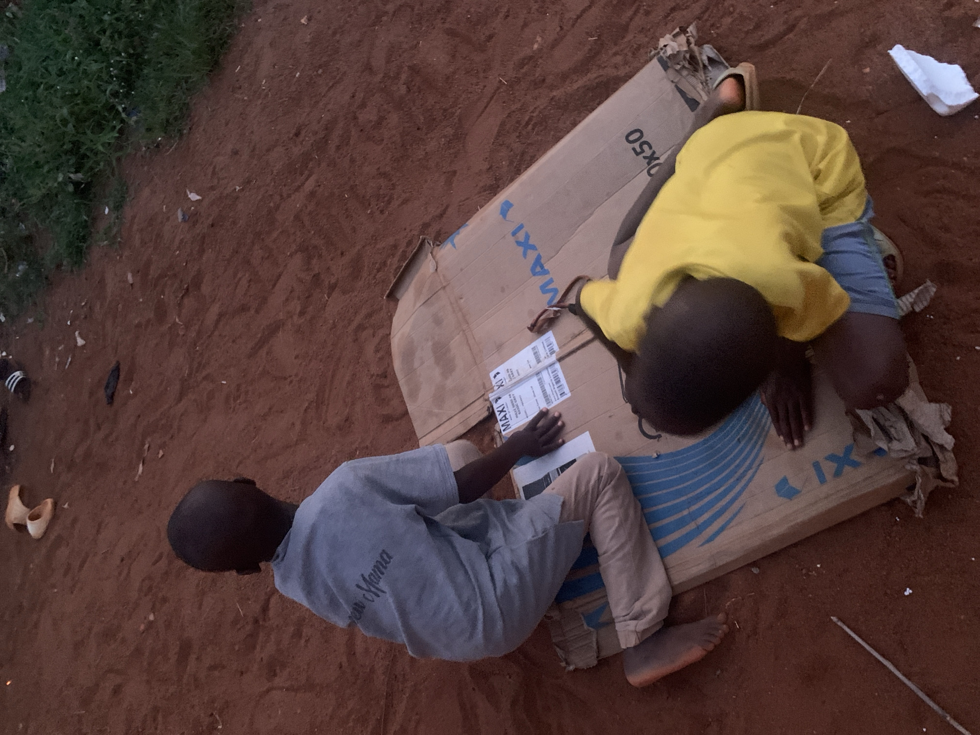 Two children are lying on a large, flat cardboard box on an unpaved surface. The box has blue and white printing on it, including the word "MAXI". One child is wearing a yellow shirt and blue shorts, the other a grey shirt and khaki pants. They appear to be interacting with something on the box, perhaps reading or drawing. The ground is reddish-brown, and there is vegetation visible in the upper left corner of the image. The location is Ukwu-Abwa, Nigeria.