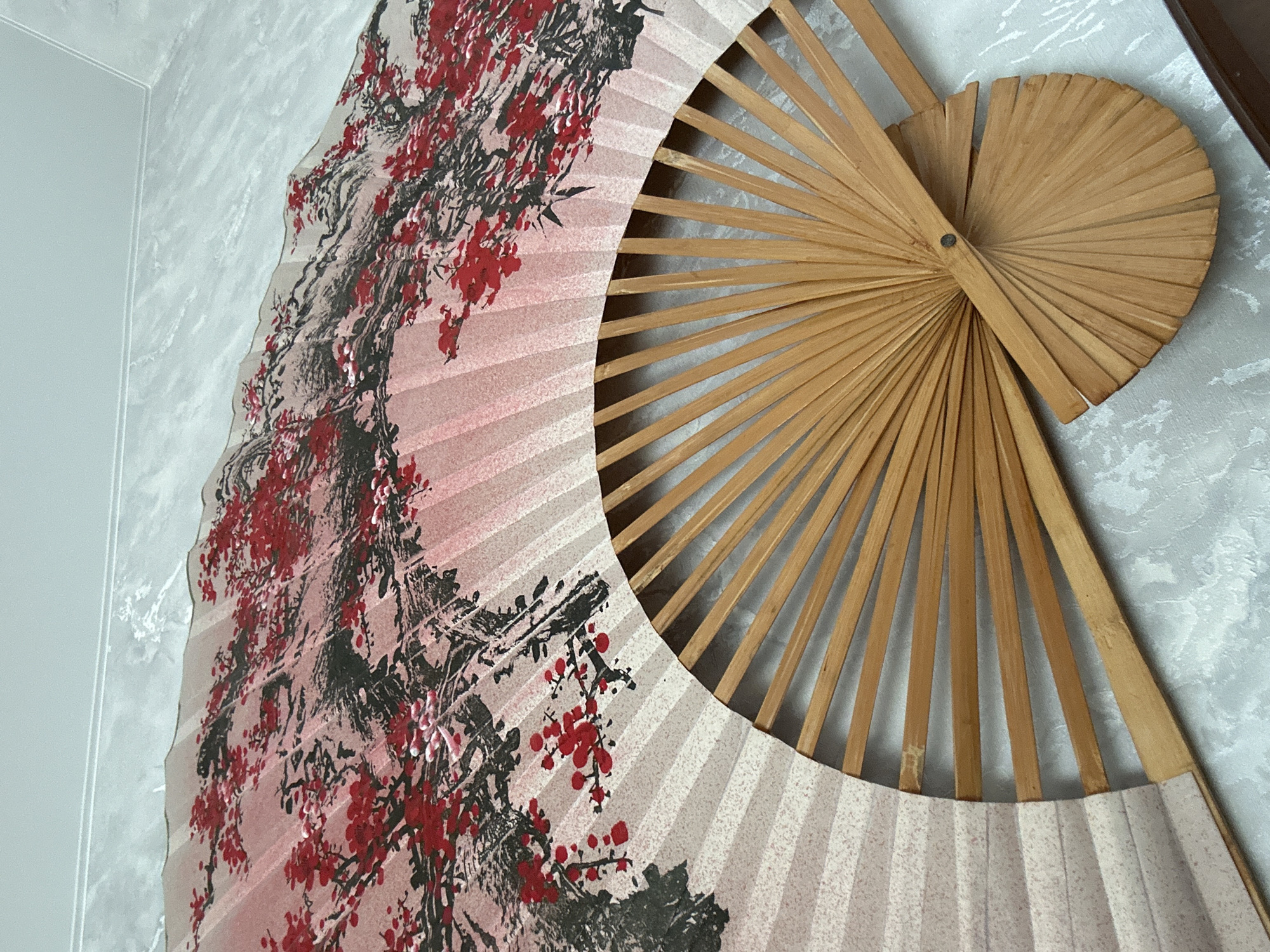 A partially opened decorative fan hangs on a light-colored wall with a textured pattern. The fan has a painted scene on its left side featuring red and black floral and branch designs. The right side of the fan displays the bare wooden struts that comprise its construction. The location is identified as Ольховка, Russia.