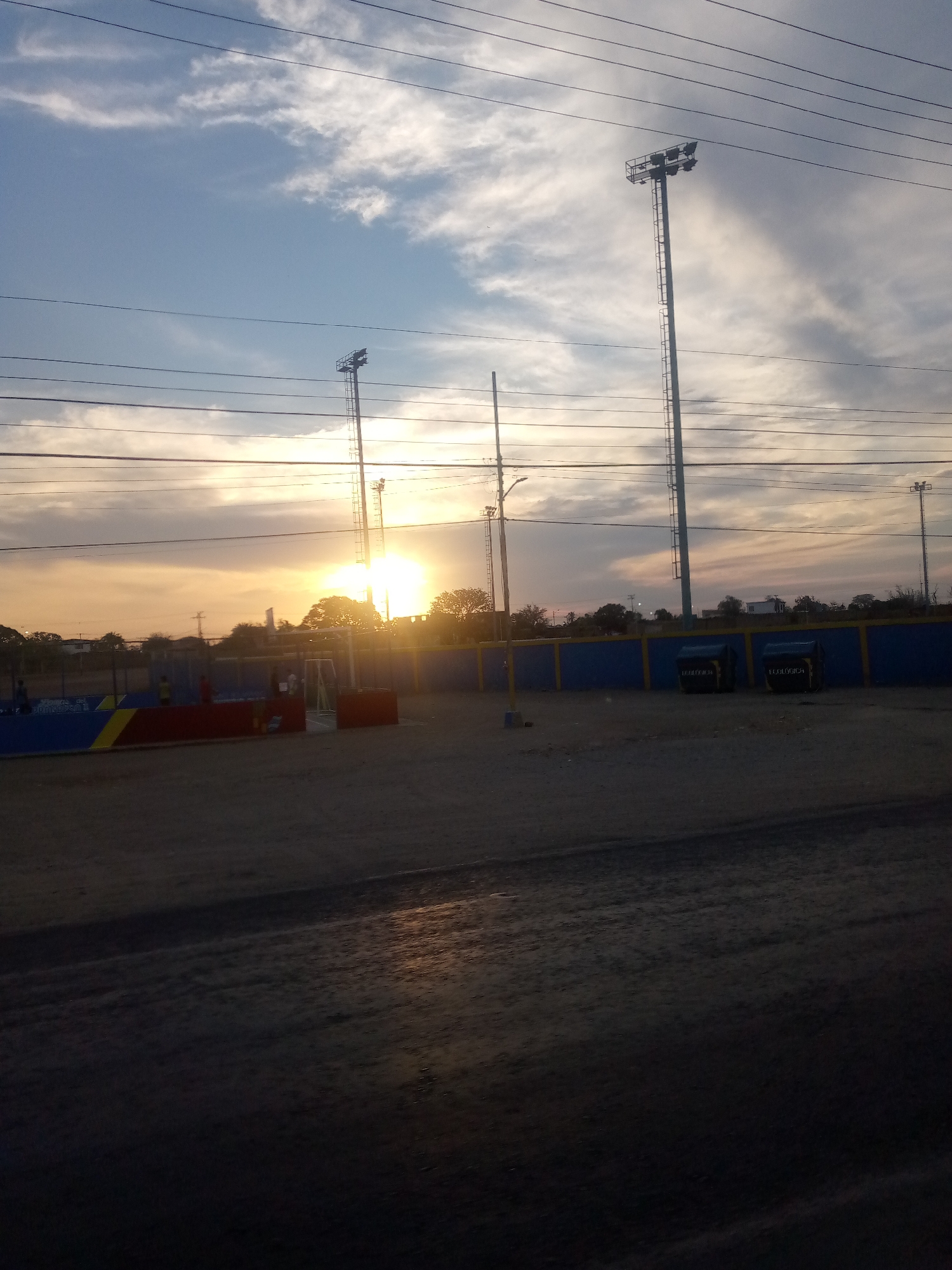 The photo shows an outdoor scene in Las Barrancas, Venezuela, during sunset. A partially visible sports field with blue and yellow barriers and a red structure is present. Lighting towers stand near the field, with a backdrop of trees and buildings silhouetted against the setting sun. Overhead power lines cross the sky. The foreground is dominated by a dark, possibly unpaved, surface. No people are clearly visible.