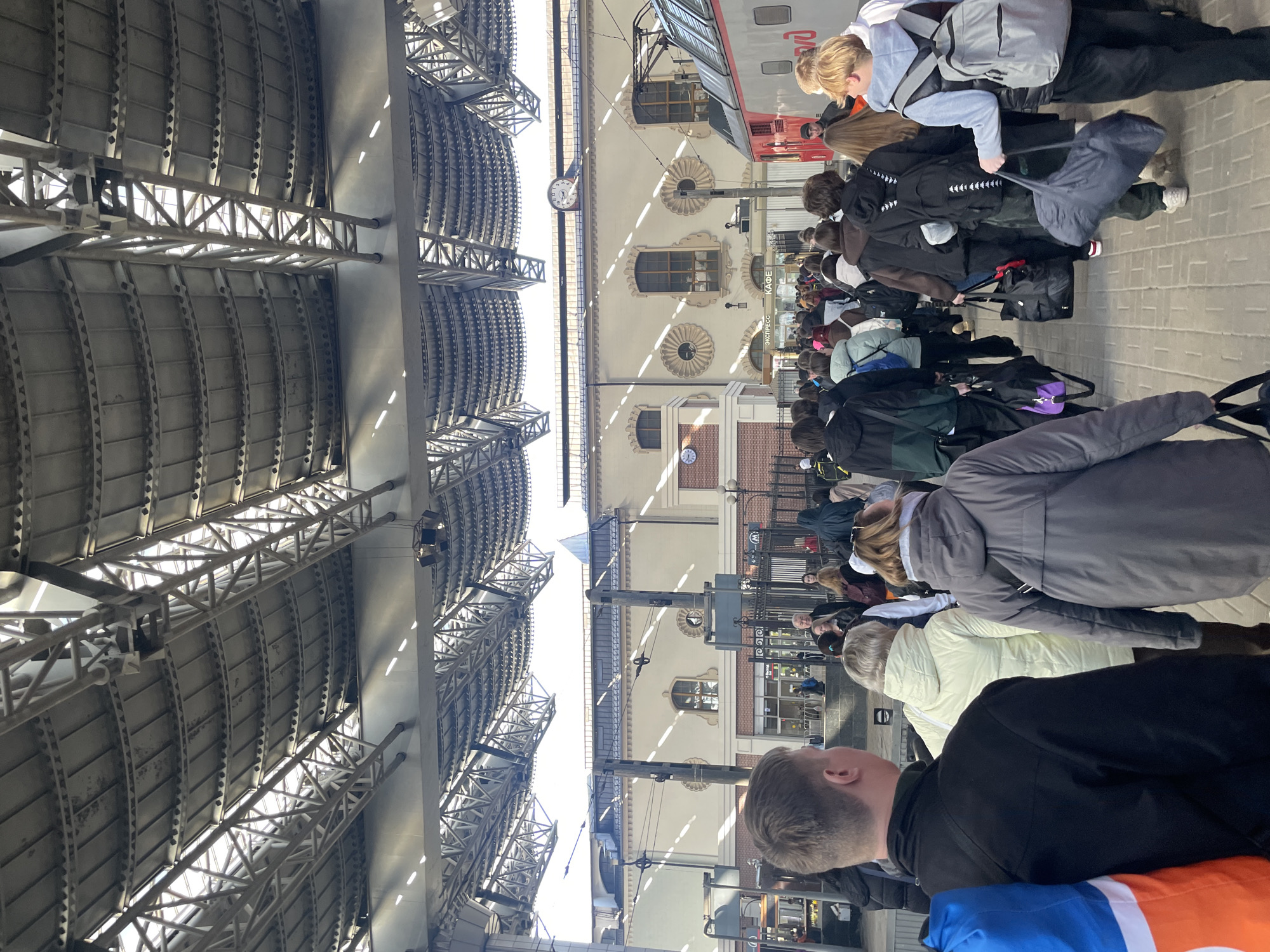 The image shows a crowded train platform at what appears to be a Moscow, Russia railway station. Numerous people with luggage are lined up alongside a train, presumably waiting to board. The station has a large, arched steel roof structure. The building adjacent to the platform is beige with decorative window surrounds. There are clocks visible on the building facade.