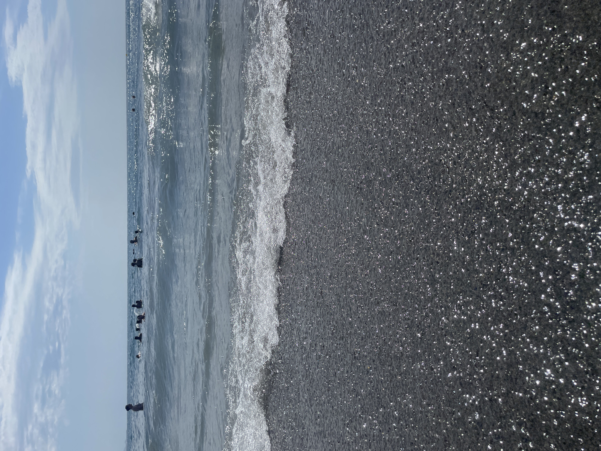 The image depicts a view of the sea and beach at the city of Almuñécar, Spain. In the background, several people are visible in the water, some appearing to be swimming or wading. The sea is calm with gentle waves, and the sunlight reflects off the surface. The foreground shows a dark, pebbly beach with numerous small, bright reflections, indicating the presence of wet sand or small stones. The sky is partly cloudy with a light blue hue.