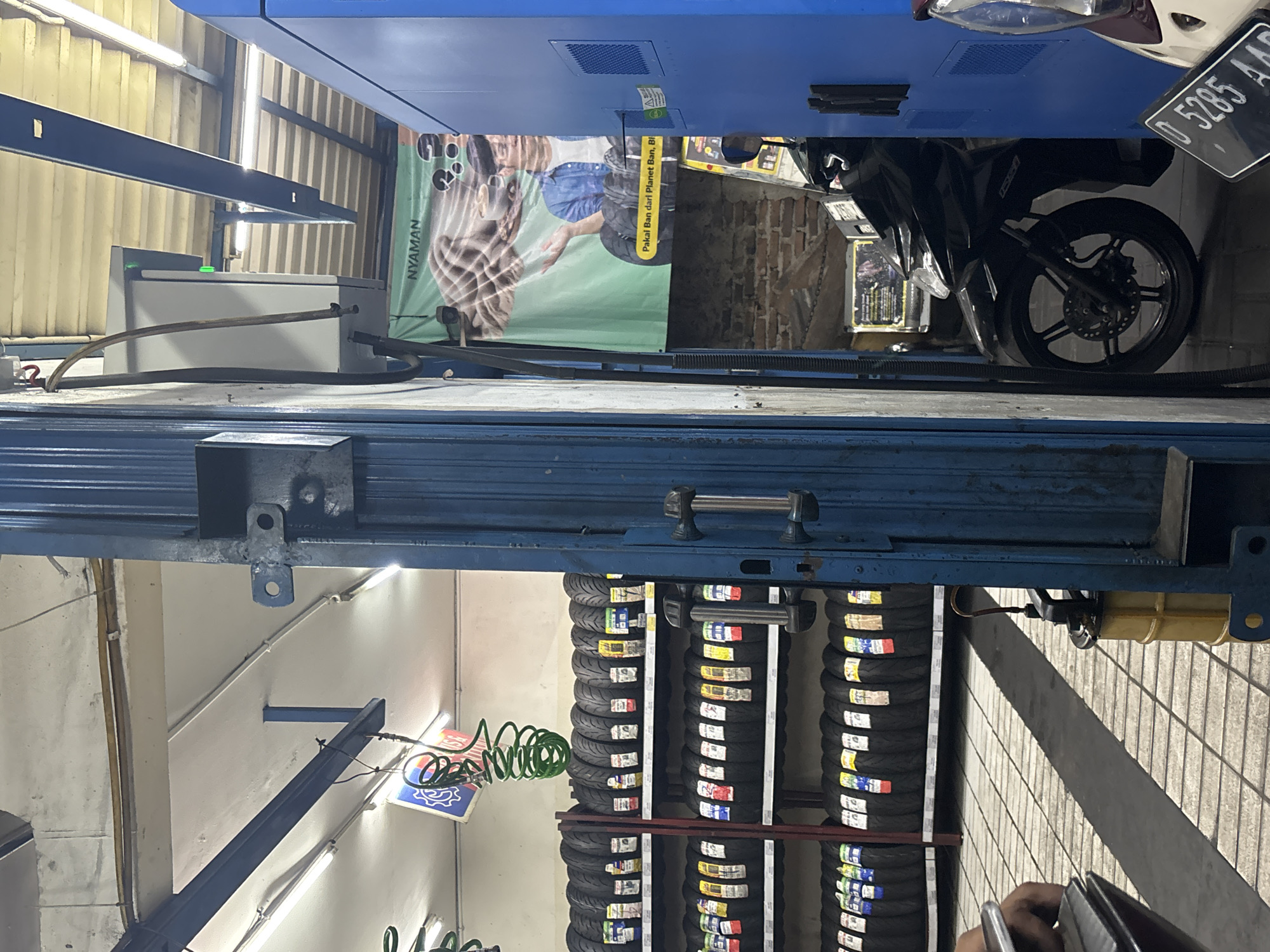 The image depicts the interior of a tire shop in North Jakarta, Indonesia, with a focus on a blue metal structure that appears to be part of a tire-changing machine or rack. Several shelves are filled with rows of new motorcycle tires, each bearing colorful price tags. To the left, a fluorescent light fixture illuminates the scene, and a coiled green hose hangs from the ceiling. A banner with an illustration of a man and coffee cups, along with Indonesian text that translates to "Comfortable," hangs on the wall in the background. A black motorcycle is parked to the right, partially obscured by the blue structure. The floor is tiled with a grey and white checkerboard pattern, and a long shadow stretches across it. A person's hand is visible in the lower left corner, holding what appears to be a clipboard or order form. A yellow oil container with a hose attached is positioned near the base of the blue structure.