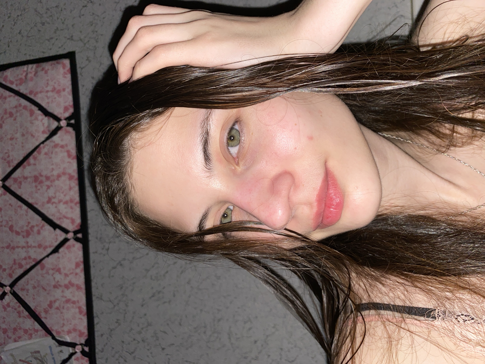 An eye-level selfie shows a young person, likely female, with fair skin, hazel-green eyes, and long, dark brown hair that appears wet. The image is rotated 90 degrees clockwise. The subject is looking directly at the camera with a calm, neutral expression, and her lips are slightly parted, revealing a natural pink color.
Her right hand is visible in the upper part of the frame, resting on or running through her wet hair near her forehead. A delicate silver chain necklace is visible around her neck, and a dark bra strap can be seen on her shoulder.
The background consists of a textured, mottled grey surface, possibly a wall, occupying most of the frame. To the far left, a section of what appears to be a decorative item is visible, featuring a pink and white speckled or patterned background with a black crisscross grid overlay. The overall lighting is dim, suggesting an indoor setting, possibly evening or night. There is no discernible text in the image. The scene is set in Votkinsk, Russia.