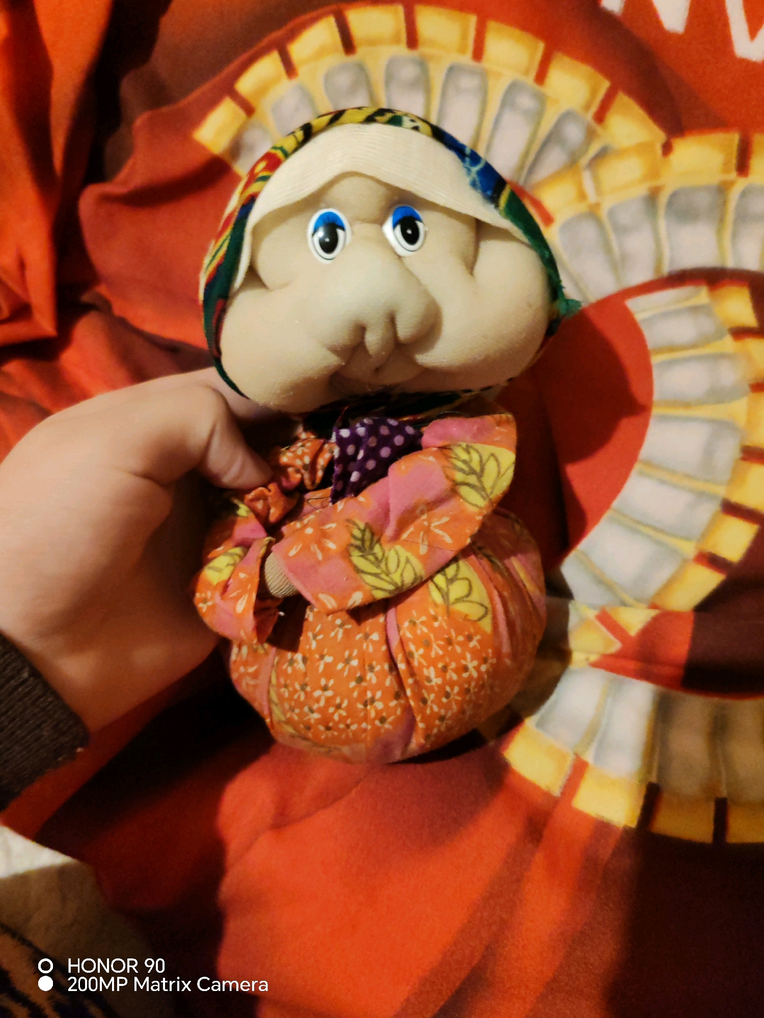 The image shows a handmade doll, likely a matryoshka doll, being held by a person's left hand. The doll has a round, pale face with blue eyes and a pinkish nose and mouth. It is wearing a colorful headscarf and a dress made of patterned fabric. The doll is positioned in front of a red and yellow patterned surface. The background is out of focus. There is a watermark at the bottom left corner that reads "HONOR 90 200MP Matrix Camera". The location in the city of Tver, Russia, is not determinable from the image.