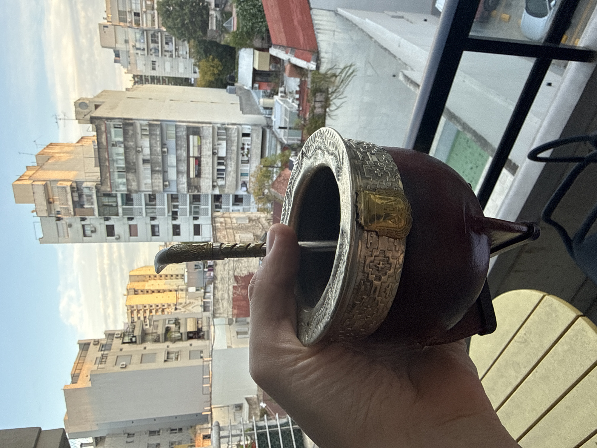 A hand holds a mate gourd with a bombilla (metal straw) on a balcony. Behind the hand and mate, an urban landscape unfolds with numerous multi-story apartment buildings, likely in Buenos Aires, Argentina, given the mate. The sky is partially visible, suggesting early morning or late afternoon light. In the immediate foreground, balcony railings and furniture are visible, further establishing the location.