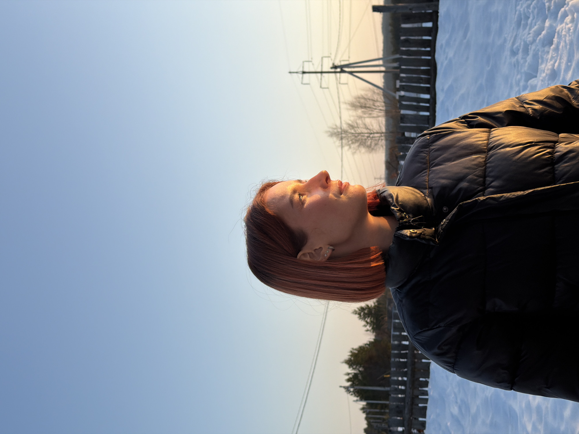 The image presents a young adult female, observed from the chest up, in profile facing right, located in Chaikovsky, Russia. She has short, straight, reddish-brown hair and is wearing a black quilted puffer jacket. Her right cheek and the right side of her jacket are illuminated by a strong, warm, golden light, indicative of either sunrise or sunset. Small silver earrings are visible in her left ear. The ground in the foreground and midground is covered in a layer of snow. In the background, a wooden fence spans horizontally, separating the immediate area from a sparse treeline. To the left, coniferous trees and utility poles with overhead wires are visible. To the right, a large metal electricity pylon stands prominently against the horizon, with additional bare deciduous trees. The sky is clear, exhibiting a gradient from light blue at the top to a pale orange-yellow hue near the horizon.