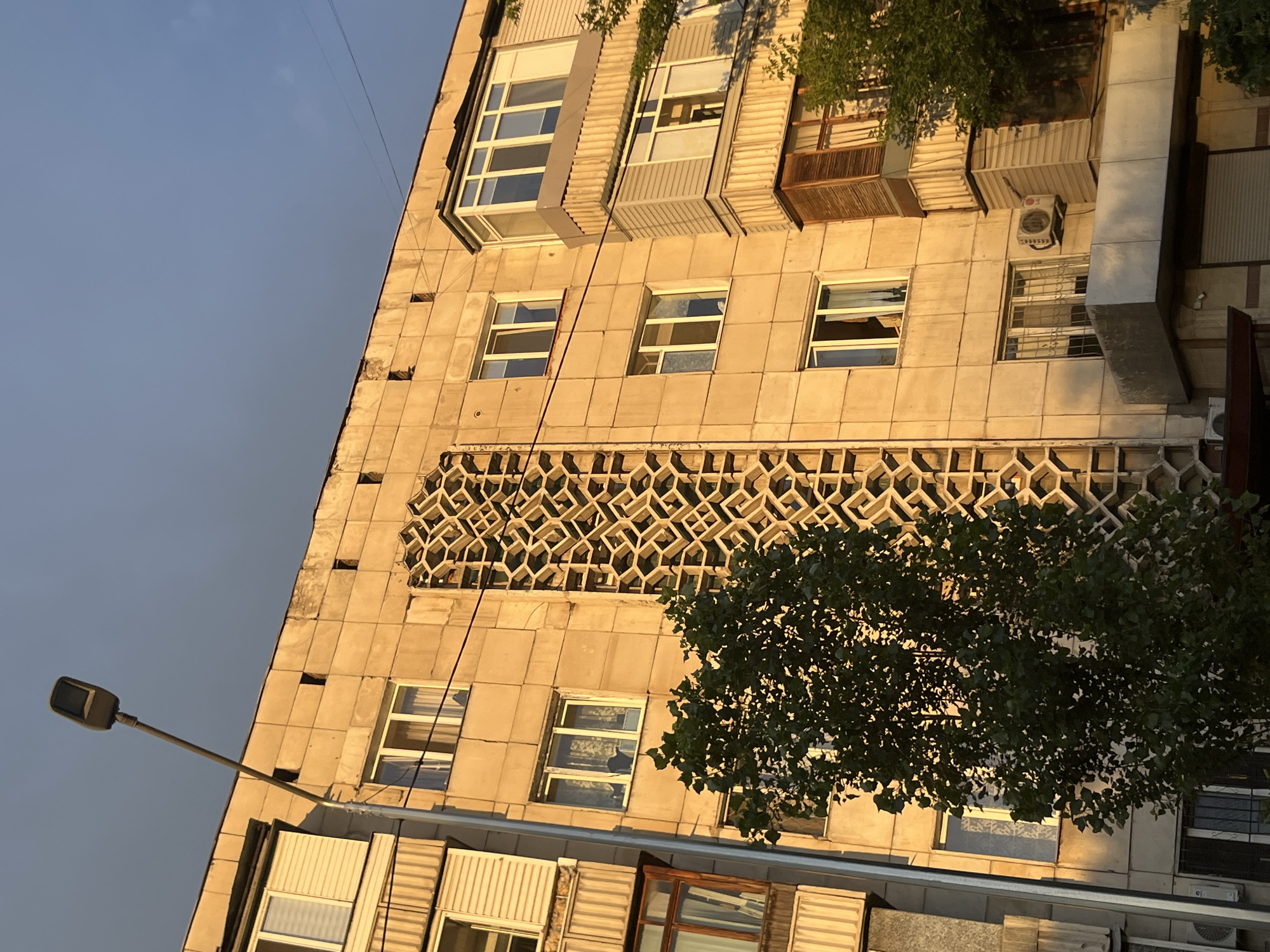 This media file shows the exterior of a building, likely an apartment complex, in Almaty, Kazakhstan. The building's facade is made of light-colored rectangular blocks and features multiple windows, some with white frames and others with darker frames. Above the main windows, there is a decorative concrete lattice pattern. Several air conditioning units are visible on the building's exterior, and a tree with lush green foliage partially obscures the facade. A street lamp is also visible on the left side of the image. The sunlight casts long shadows, suggesting it is either early morning or late afternoon. The sky is clear and blue, indicating fair weather. The architecture appears to be from the Soviet era, common in many cities of Kazakhstan. No people are visible in the frame, and there is no discernible text. The scene evokes a sense of everyday urban life in a post-Soviet city.