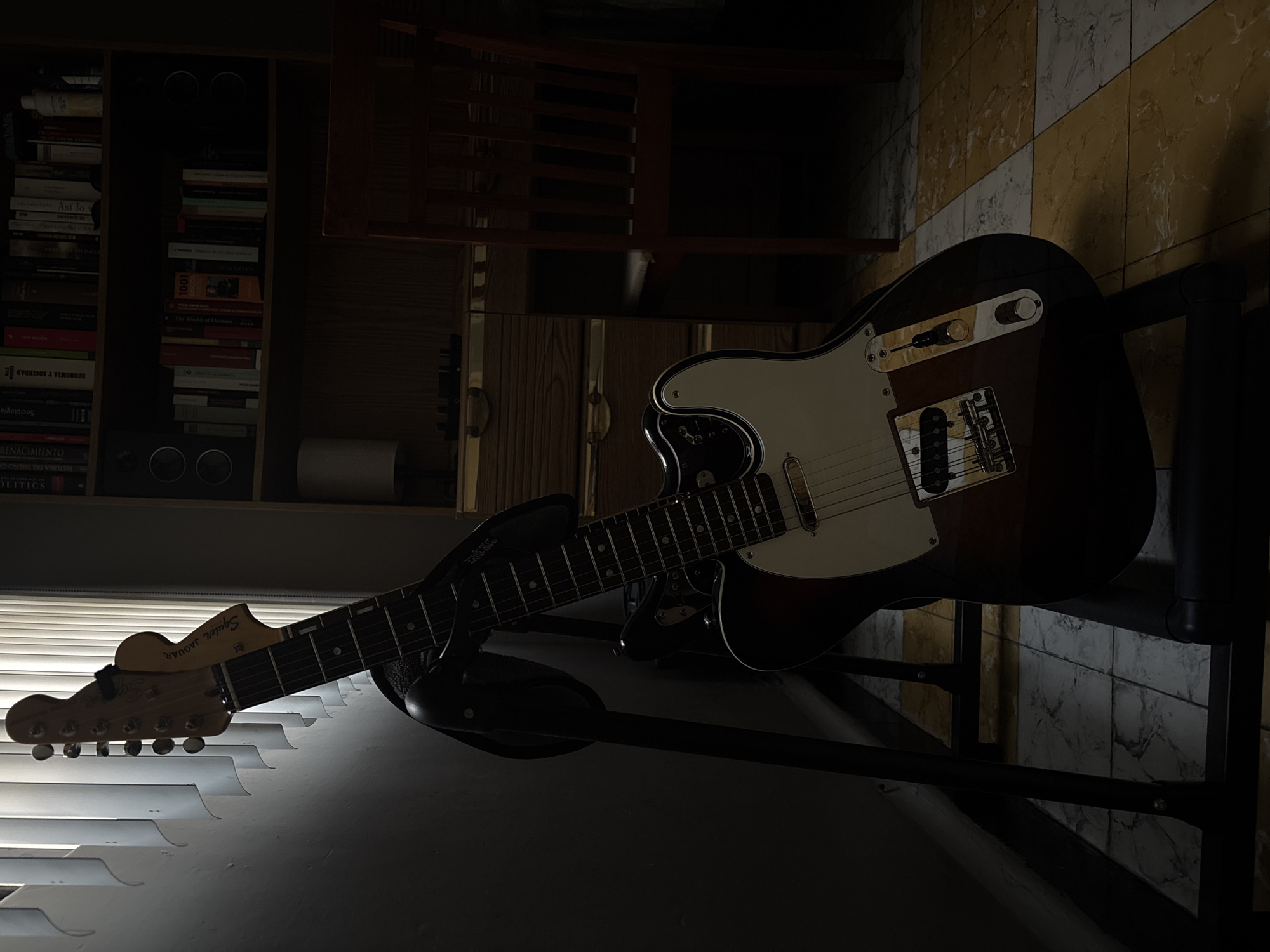 The image features a Squier Jaguar electric guitar resting on a stand in a dimly lit room. The guitar has a white pickguard and a dark brown body. Behind the guitar, there's a bookshelf filled with books, some of whose spines are visible and readable. A chair is also situated behind the guitar. The wall behind the guitar and chair is tiled in a yellow and white pattern. Blinds are visible in the foreground, suggesting a window. The image's orientation is skewed, with the guitar presented diagonally across the frame. There's no indication of Puebla, Mexico, in the image.