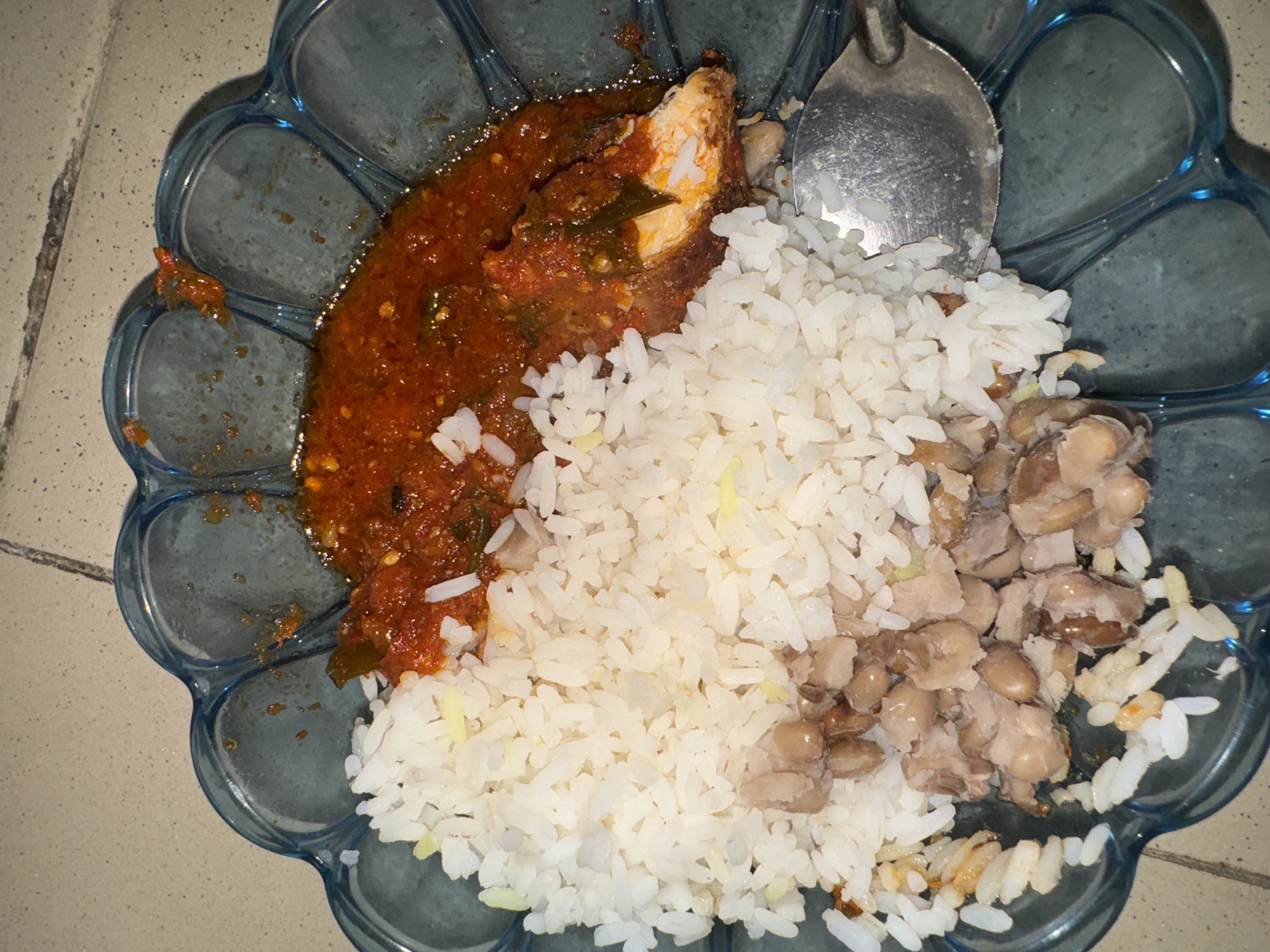 A plate of food is placed on a surface. The plate is blue with a decorative, scalloped edge. The food consists of a portion of rice, beans, and a reddish sauce with what appears to be fish. A silver spoon rests on top of the rice. The surface beneath the plate appears to be tiled. There is no visible indication of location or city. The city Sabon Tasha, Nigeria is not visibly present in the image.