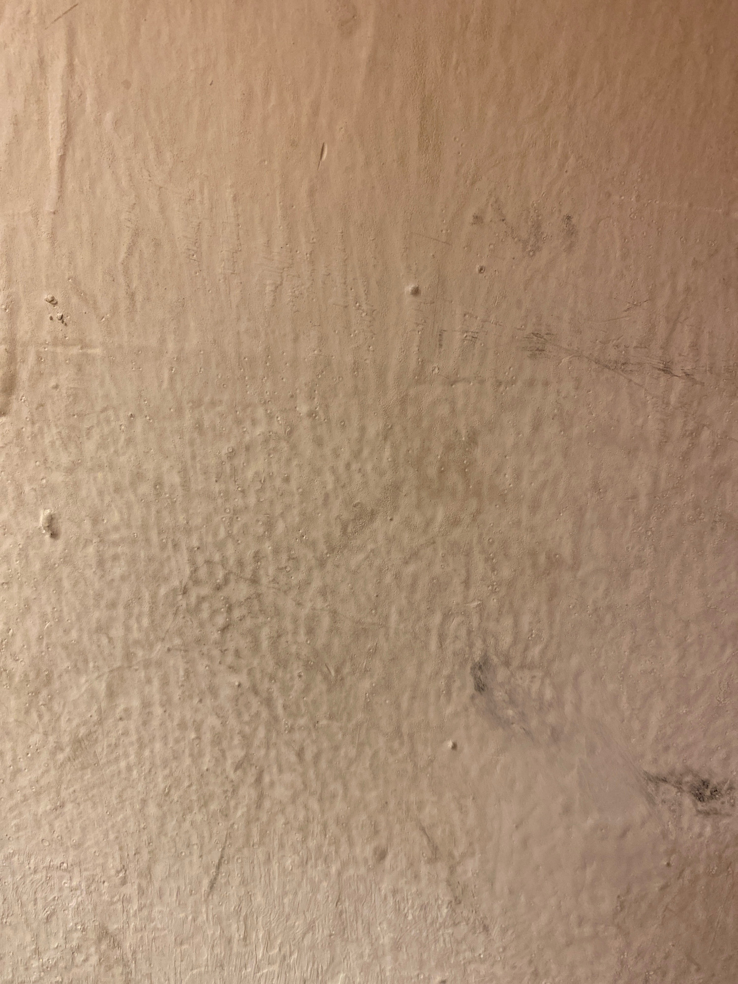 The image shows a close-up of a textured, beige-colored wall. The surface is uneven, with subtle bumps and indentations, possibly indicating a stucco or plaster finish. There are some faint lines and smudges visible, suggesting wear or age. The lighting appears to be artificial, casting a warm, soft glow on the wall. No people, objects, or specific environmental features are discernible that would allow for identification of a location within Sankt Johann im Pongau, Austria. The image is purely a textural study of a wall surface.
