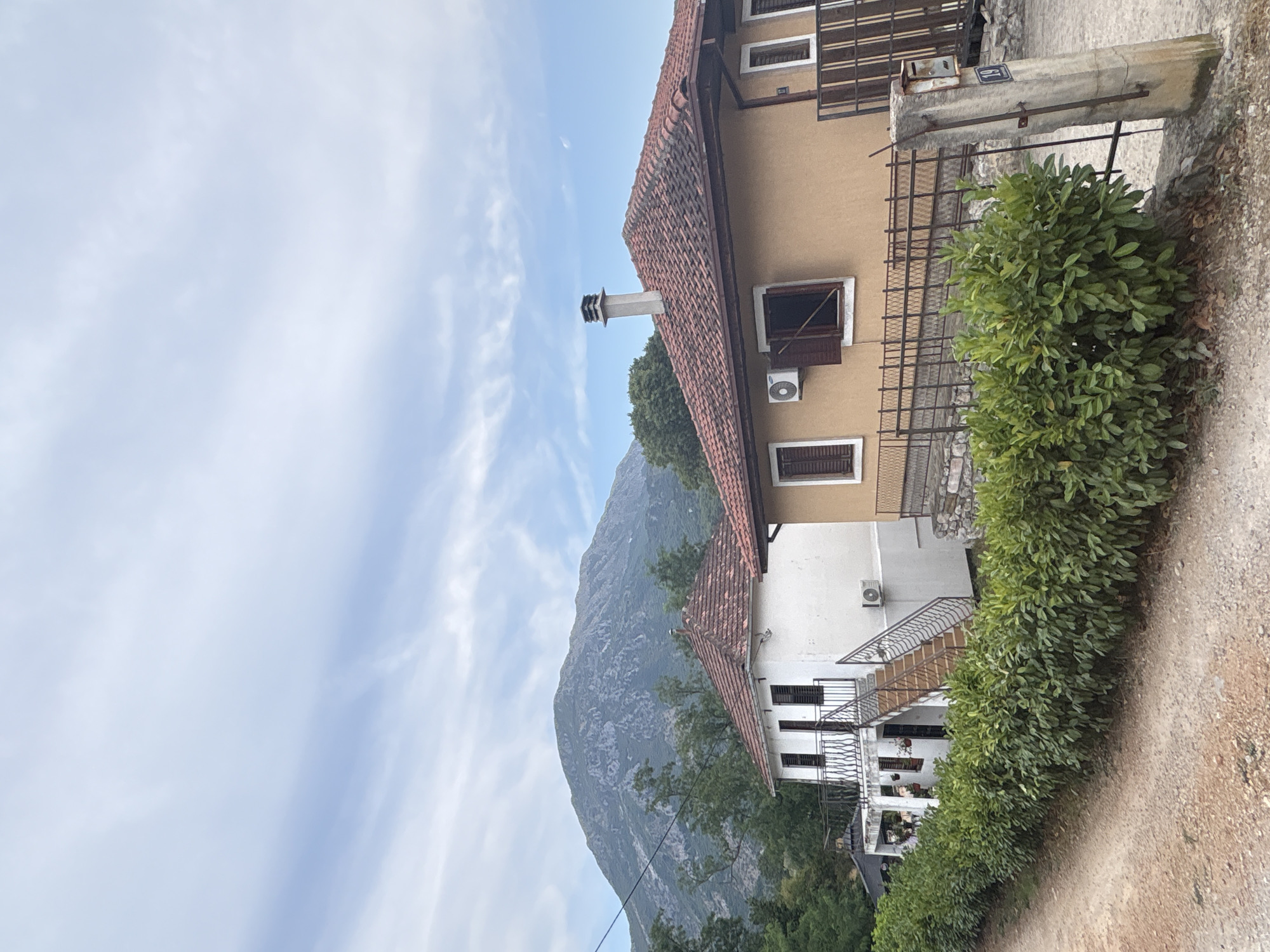 The image depicts a rural scene in Stari Bar, Montenegro, featuring two houses nestled against a backdrop of a large, rocky mountain under a cloudy sky. The foreground is dominated by a dirt road on the left and a well-maintained green hedge bordering the properties.
On the left side of the image is a white, two-story house with a red tile roof. It has multiple windows with dark shutters and a balcony on the upper level accessible by an external staircase. An air conditioning unit is visible on the lower part of the wall. Flower pots are placed on the balcony.
To the right of the white house is a tan-colored, single-story house, also with a red tile roof. This house has two windows, one with wooden shutters partially open. A modern chimney extends from the roof. An air conditioning unit is mounted on the wall near one of the windows. A fence with a metal railing separates the property from the dirt road. A concrete post with a mailbox and a sign indicating the number "19" stands at the edge of the property.
The mountain in the background is imposing, with visible rock formations and sparse vegetation. Trees are scattered around the base of the mountain and between the houses, suggesting a natural, somewhat undeveloped environment. The sky is partly cloudy, with streaks of white clouds against a pale blue backdrop. The overall impression is one of a peaceful, rural setting.