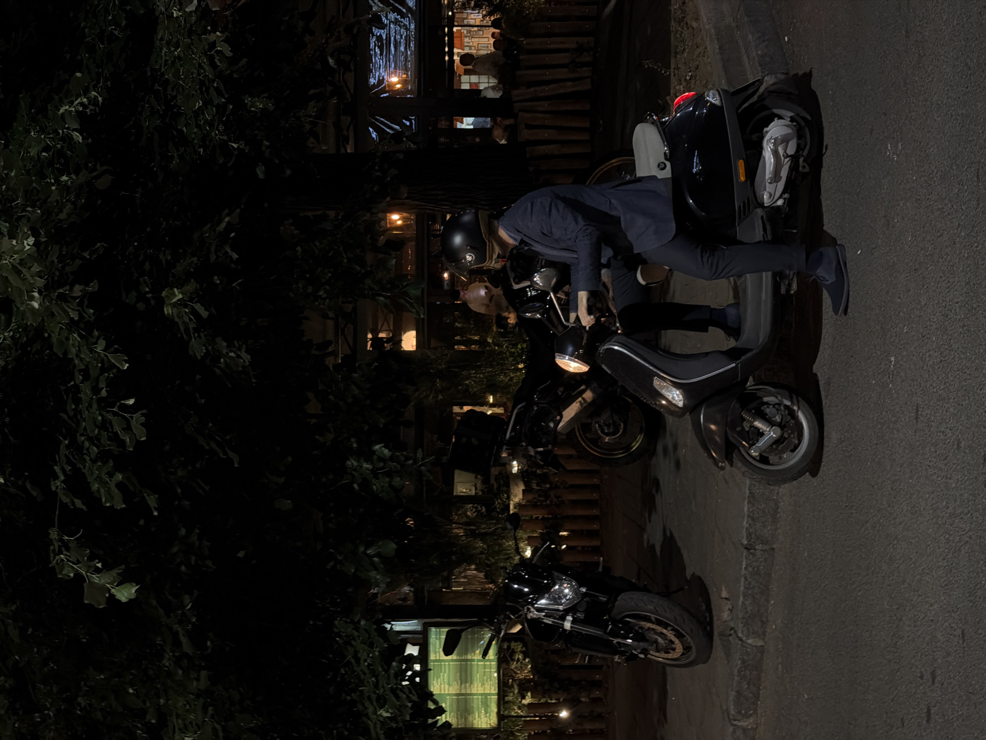 The image shows a man in a helmet getting on a black scooter on a street in Vracar Urban Municipality, Serbia, at night. There are two other motorcycles parked nearby. A building with lights and trees is in the background. The street is paved, and a sidewalk borders it.