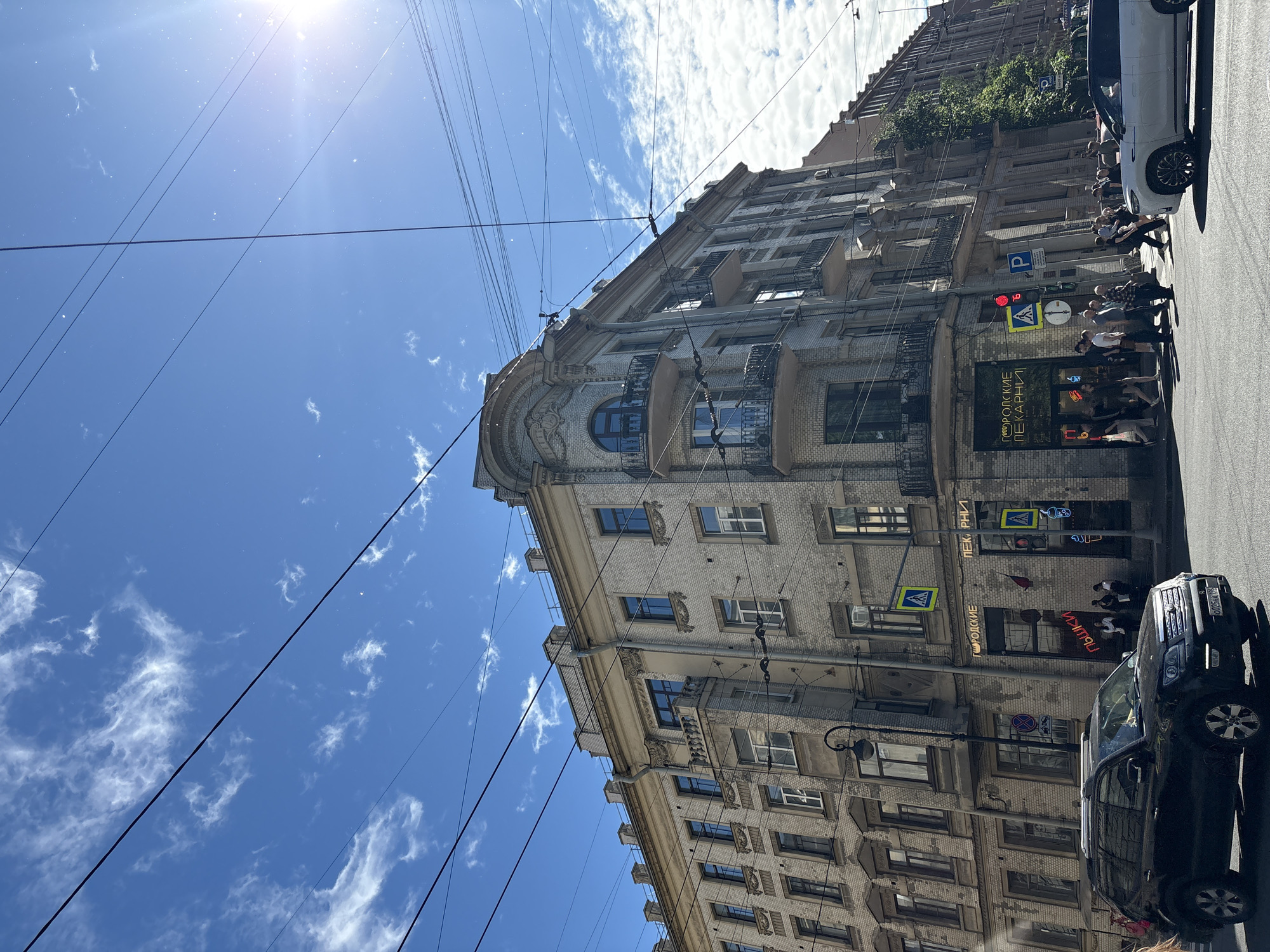 In Saint Petersburg, Russia, on a bright, cloudy day, a multi-story building with ornate architectural details stands prominently on a street corner. The building, constructed of light-colored brick, features numerous windows, balconies with wrought-iron railings, and decorative moldings. The facade exhibits signs for "ГОРОДСКИЕ ПЕКАРНИ" (City Bakeries) and "ПЫШКИ" (Pyshki), indicating commercial establishments on the ground floor.
The street in front of the building is a busy intersection with vehicular traffic and pedestrians. A black SUV is parked on the left side of the road, and a white SUV is moving on the right. Pedestrians are crossing the street at a designated crosswalk, with traffic lights visible. Overhead, a network of electrical wires and tram lines crisscrosses the sky, a common feature in the city. The bright sun casts a glare on the scene, with scattered white clouds adding texture to the blue sky. The overall impression is a snapshot of urban life in a historic European city.