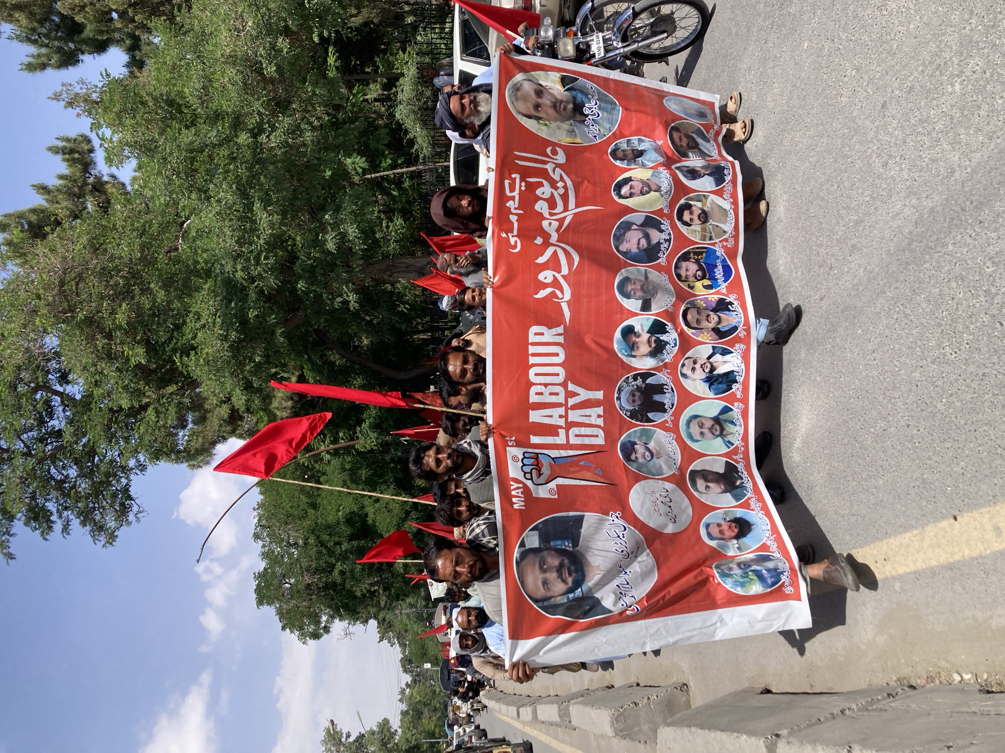 A group of men are holding a large banner for Labour Day in Quetta City Tehsil, Pakistan. The banner is red and white, with photos of men in circles along its surface. It also has writing in English and Arabic. Several people are holding up red flags near the banner. The scene is outdoors, and there is a road and greenery in the background. A motorcycle is partially visible in the upper right corner of the frame.