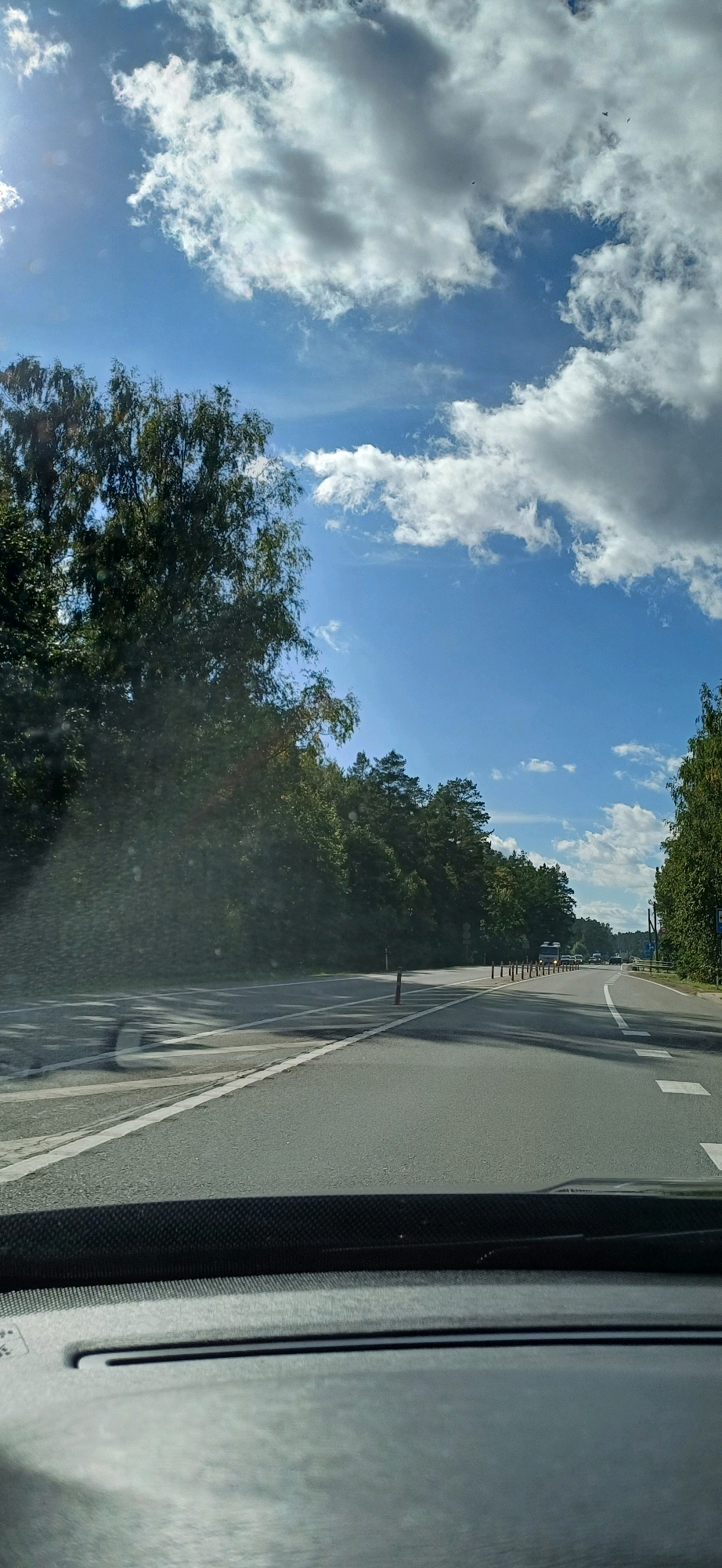 The image shows a view from inside a vehicle looking out at a road surrounded by trees. The road appears to be a highway or a similar multi-lane thoroughfare. There are white lane markings on the asphalt, and a line of bollards separates lanes. The trees on either side are dense and green, indicating a natural, possibly forested, environment. The sky above is blue with scattered white clouds, suggesting a sunny day. There are a few vehicles visible in the distance on the road, including a truck. The dashboard of the vehicle is visible in the foreground, partially obscuring the view. The overall scene suggests a road trip or travel through a scenic, wooded area.