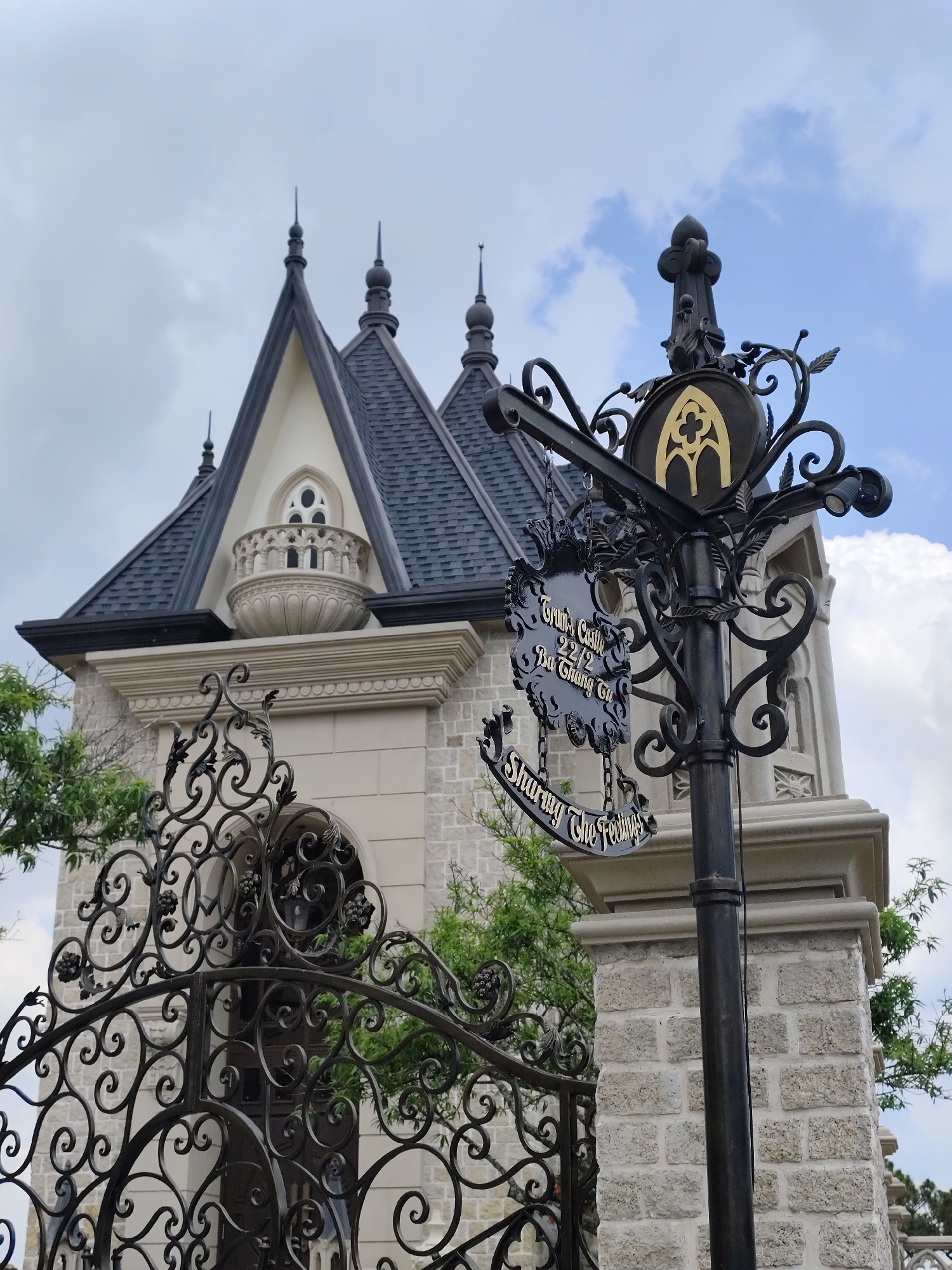 The image captures a view of "Trum's Castle" located at 22/2 Ba Trung in Da Lat, Vietnam. The building, visible in the background, features a distinct architectural style with pointed roofs and a balcony, constructed from light-colored stone. An ornate wrought-iron gate and a decorative signpost are prominently featured. The signpost, with black and gold accents, bears the castle's name and a tagline that reads "Sharing The Feelings." The sky is overcast with patchy clouds. Green foliage peeks through the gate and around the signpost, adding a touch of nature to the scene. The overall impression is of an upscale, possibly European-inspired establishment in a Vietnamese setting.