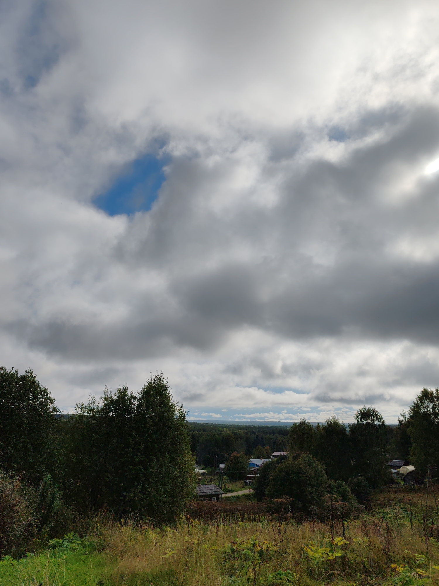 This is a photograph taken from an elevated vantage point overlooking a rural landscape. In the foreground, there is tall, dry grass and some green vegetation. Behind this, a cluster of trees dominates the middle ground, with a small village nestled amongst them. The village consists of several buildings with dark roofs, some appearing to be houses and others possibly agricultural structures like greenhouses. Beyond the village, a dense forest stretches to the horizon. The sky is filled with dramatic, heavy clouds, with patches of blue sky visible. The overall impression is of a somewhat overcast, late summer or early autumn day in a remote, rustic setting. The location is identified as Chukhlem, Russia. There are no people or animals visible in the image, and no discernable actions or interactions are taking place.
