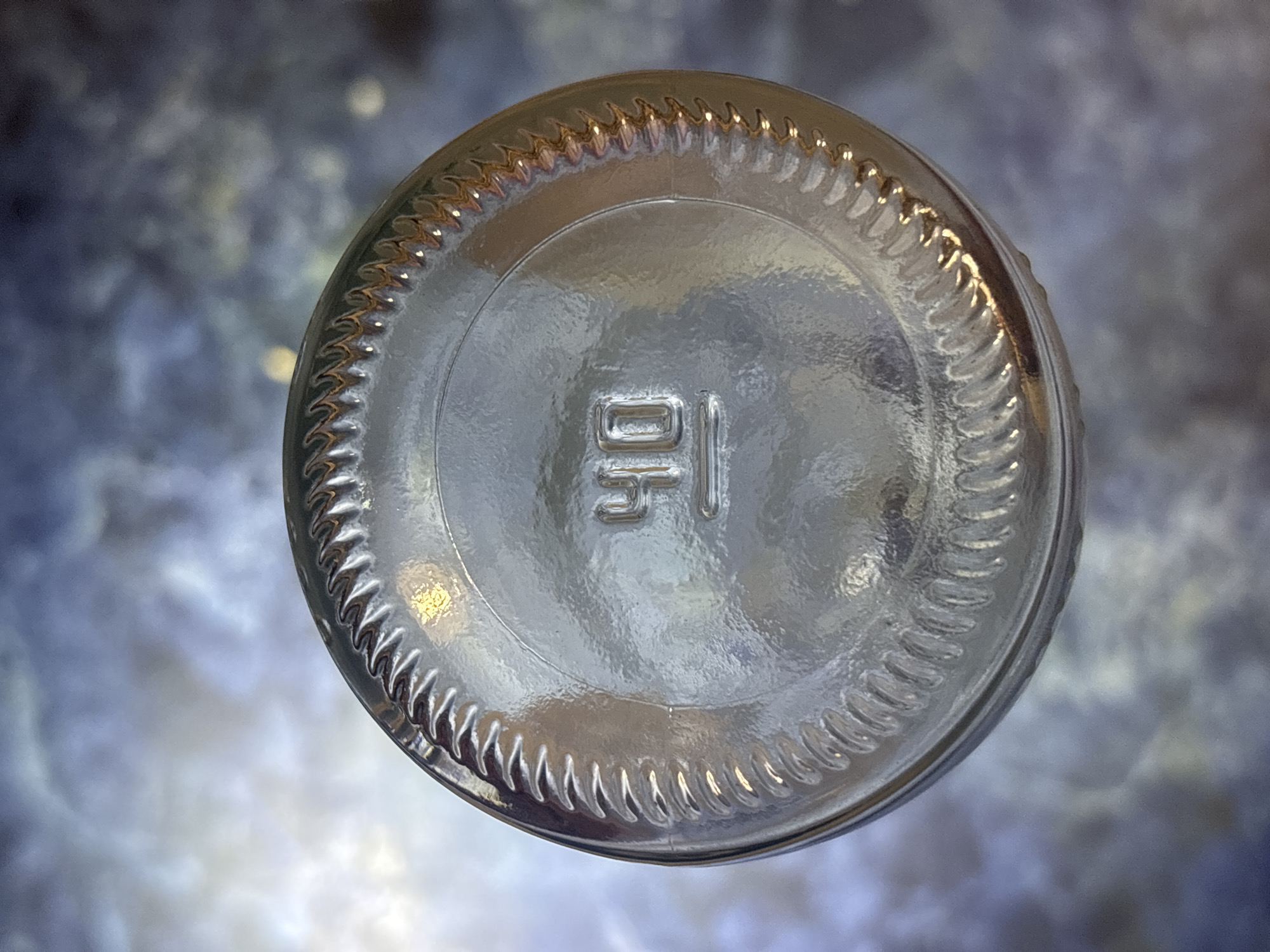 The image shows the bottom of a clear glass bottle with a ridged edge and the number "40" embossed in the center. The bottle rests on a surface with a mottled, bluish-gray and white pattern, which could be a textured countertop or a fabric. There are some reflections and light sources visible on the bottle, suggesting it is illuminated from above and to the side. The background is out of focus and displays a similar mottled pattern. The scene is indoors, and there is no information available to determine its specific location in the city of Kremenchuk, Ukraine. There are no people, actions, or interactions depicted in the image.