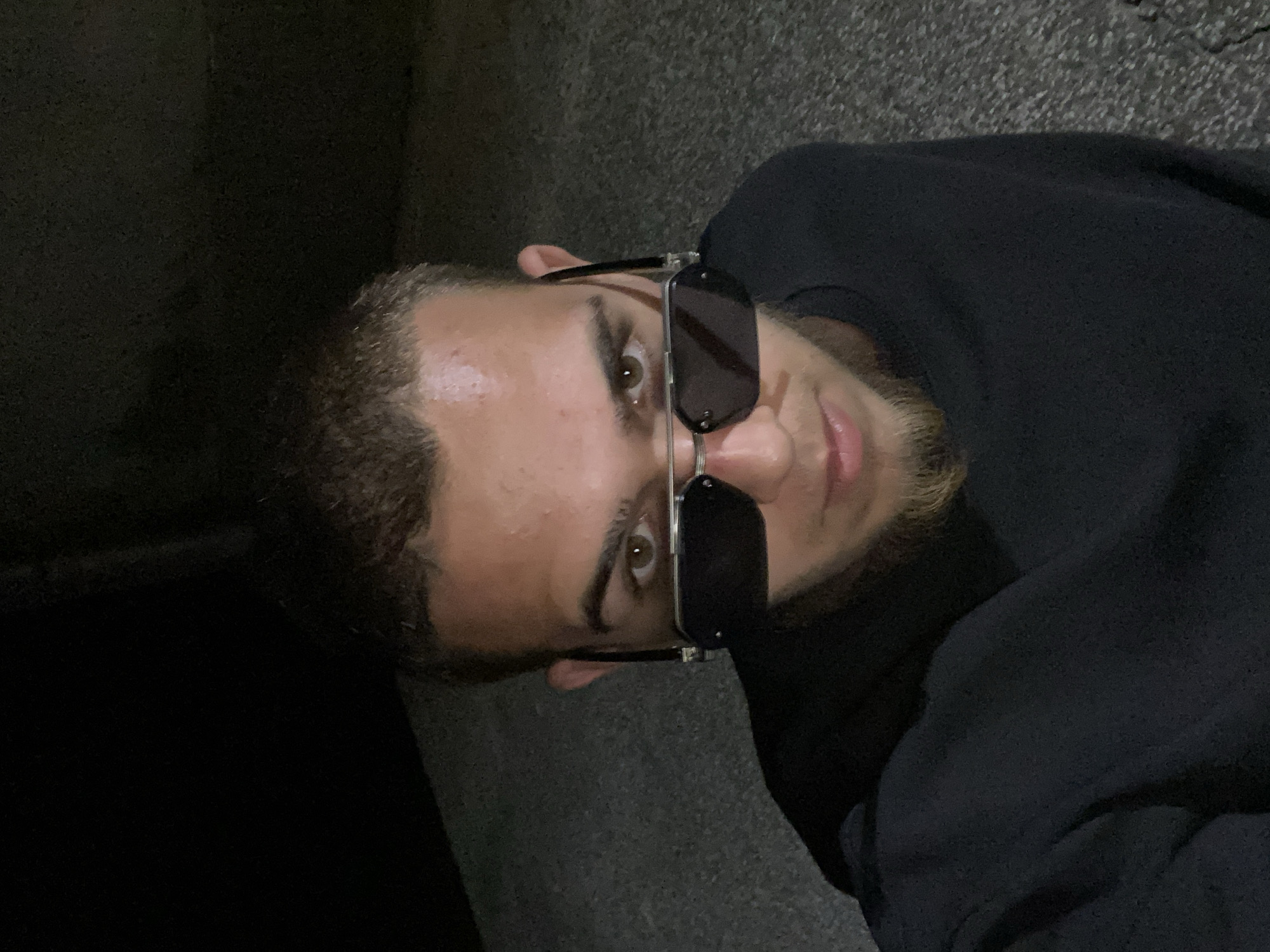 An individual is captured in a low-angle, front-facing selfie, their face partially obscured by dark, rectangular sunglasses with silver frames. They have short, light brown hair and a goatee. The subject is wearing a dark, long-sleeved shirt. The background appears to be an outdoor, urban environment, characterized by a rough, asphalt-like texture on the ground. The lighting suggests it is nighttime or a dimly lit area. The location is specified as Hirevo, Bulgaria.