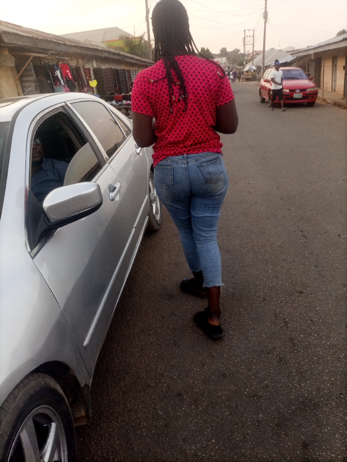 A person in a red polka dot shirt and jeans walks beside a silver car on an asphalt road. The person's back is to the camera. In the driver's seat of the car, an individual wearing sunglasses can be seen. A red car and figures are visible in the distance. Buildings line the street. The overall scene appears to be outdoors during the day.