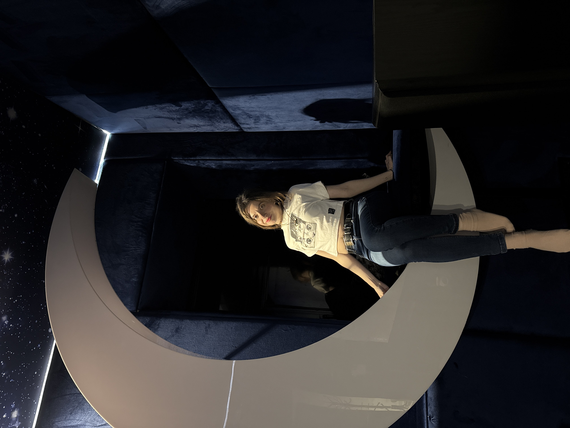 A woman is lying in a white crescent moon prop. She has short brown hair and is wearing a white shirt and dark wash jeans with socks. The background is an exhibit in a dark room with a star-speckled wall in Moscow, Russia.