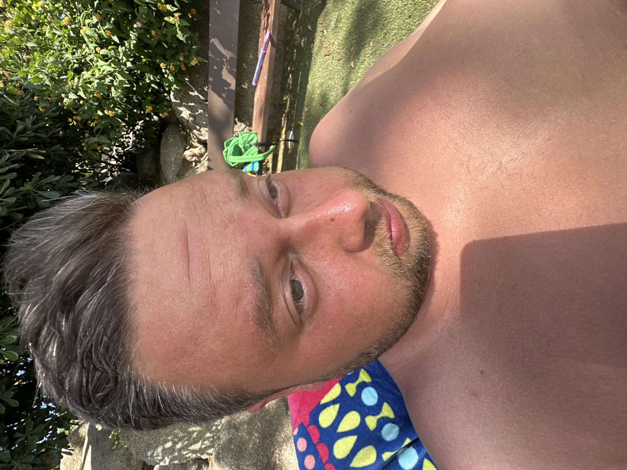 The image shows a close-up, overhead shot of a man with light skin and thinning brown hair, who appears to be sunbathing outdoors. He is partially visible, from the chest up, with his head tilted and eyes looking to the left. His skin is flushed and appears to have goosebumps in places. He is lying on a colorful towel with a pattern of blue, pink, yellow, and white shapes. In the background, there is green foliage, rocks, and a wooden bench with a bright green object and a pen on it. The scene suggests an outdoor setting, possibly a garden or poolside area, consistent with the climate of Sant Feliu de Guíxols, Spain, which is known for its coastal location and Mediterranean vegetation. The lighting indicates a sunny day.