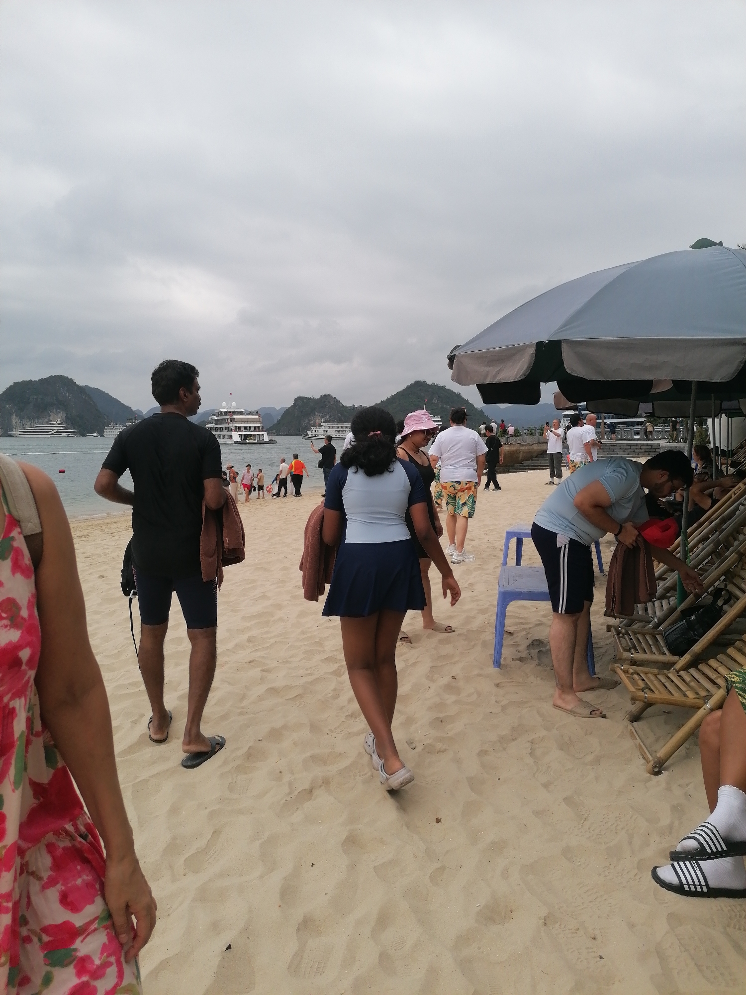 The image depicts a lively beach scene on an overcast day, likely in Bai Chay Ward, Vietnam. The foreground is dominated by light-colored sand, showing footprints and several individuals.
On the left, a person in a floral dress is partially visible. Centrally, a man in a black t-shirt and dark shorts, holding a brown towel and carrying a backpack, walks away from the camera towards the water. Adjacent to him, a woman in a dark blue skirted swimsuit and light blue top also heads towards the water. To the right, a man in a light blue t-shirt and dark shorts bends over a set of bamboo beach chairs, holding a red hat, seemingly organizing items beneath a large grey and green beach umbrella. A blue plastic stool is positioned nearby.
In the middle ground, more people are seen walking and gathering on the beach near the water's edge. The background features calm, greyish-blue water where several large, multi-deck boats or cruise ships are anchored or moving, accompanied by smaller vessels. Beyond the water, dramatic limestone karsts or mountains, characteristic of coastal Vietnam, rise prominently under a uniformly cloudy sky.
The weather appears mild due to the overcast conditions. People are dressed casually in beach attire, including swimwear, shorts, and t-shirts; some are barefoot, while others wear flip-flops or slides. The overall atmosphere suggests a leisurely day at a popular tourist beach. No legible text is visible within the frame.