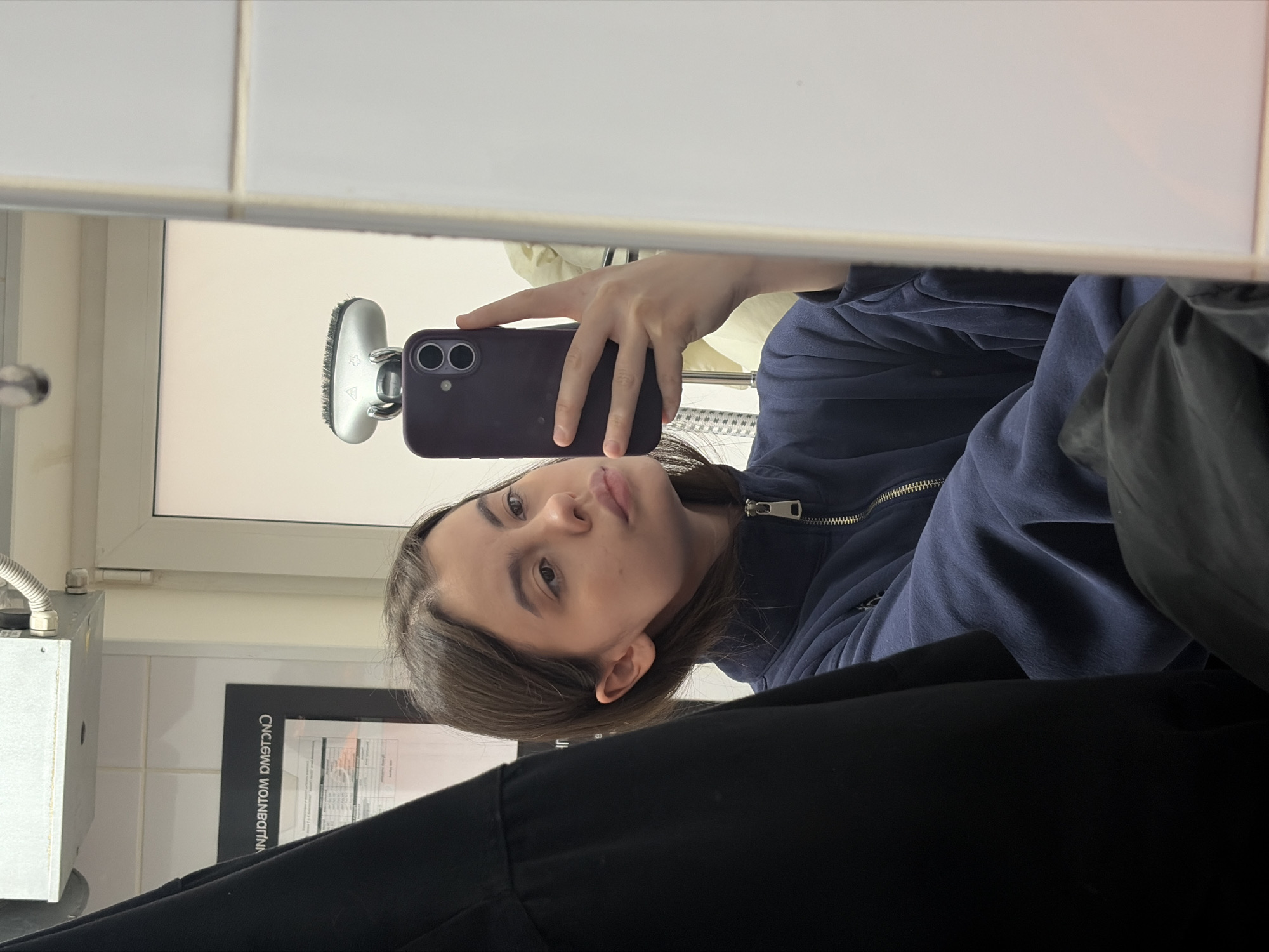 The image is a mirror selfie taken by a young woman with brown hair. She's wearing a blue zippered jacket and a black garment is partially visible in the foreground. She is holding a purple smartphone in her right hand, capturing the image. The reflection reveals a bathroom setting with white tiled walls and an open window in the background. There is a hair-washing station with a silver faucet and a black information poster on the wall to the left. The location cannot be determined to be in the city of Chaikovsky, Russia from the image.