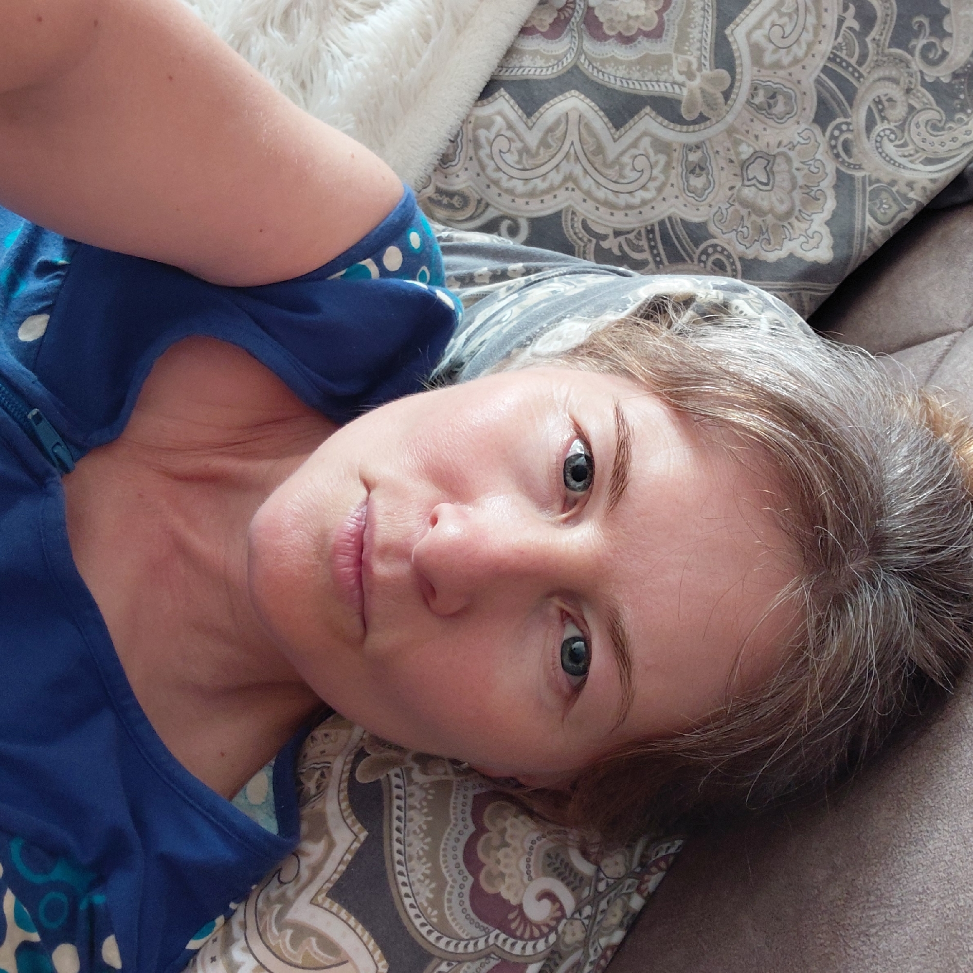 A medium shot, taken from above, shows a fair-skinned woman with blonde and graying hair pulled up in a bun, lying on a sofa. She is looking directly at the camera with a neutral expression. Her eyes are blue-green, and she has fair skin with some visible pores and fine lines. She is wearing a blue V-neck top with a zipper closure and a white and teal floral pattern. The background consists of patterned pillows with a paisley design in gray, cream, and burgundy, and a textured brown sofa. A fluffy white blanket is visible to the left. The overall lighting is bright and even, suggesting it is daytime, possibly near a window. The scene does not contain any identifying landmarks or features that would indicate a specific location within Novosibirsk, Russia, or any other city.
